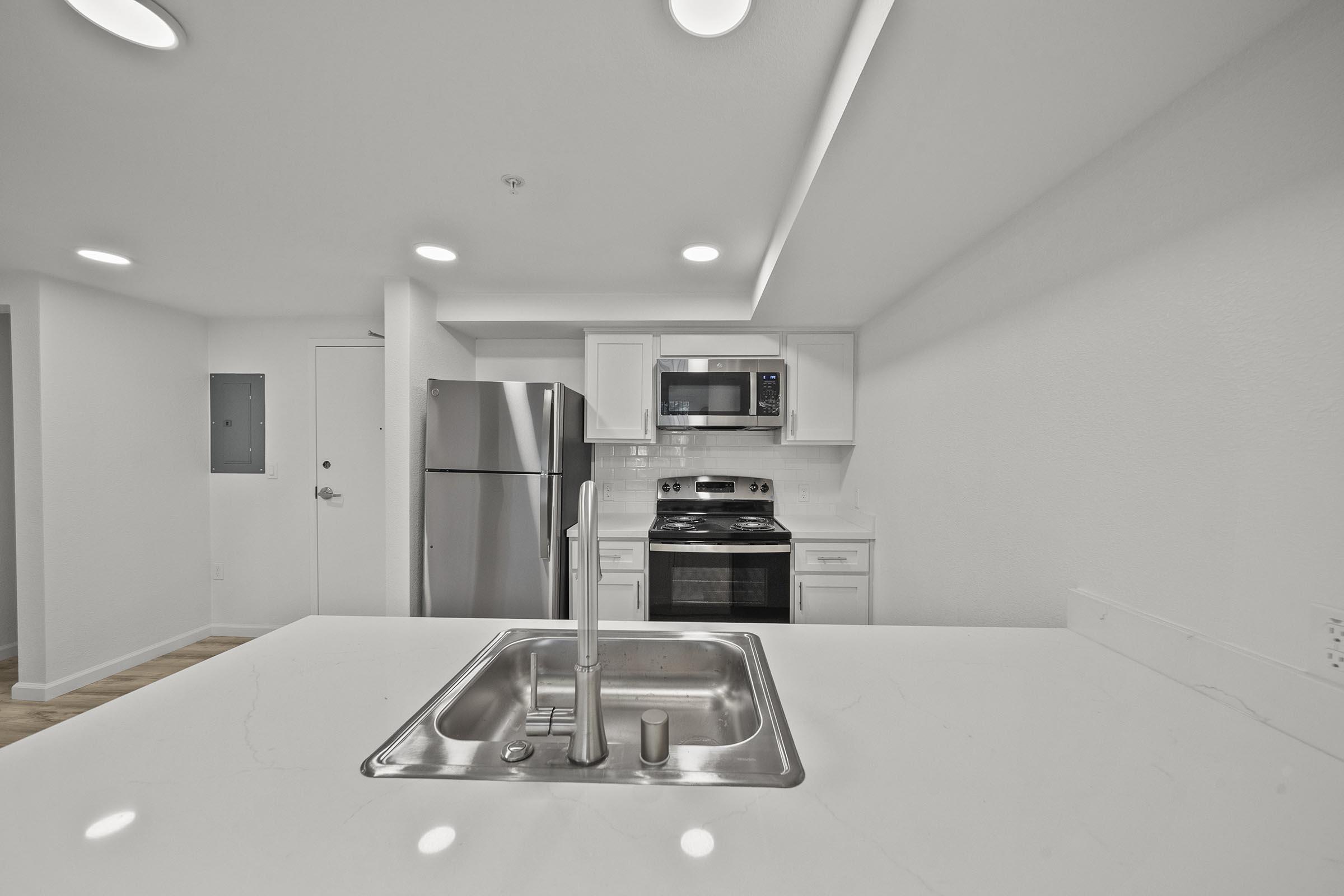 Modern kitchen with a central island featuring a sink. Stainless steel appliances, including a refrigerator and an oven, are visible in white cabinetry. The space is well-lit with recessed ceiling lights and has a clean, minimalist aesthetic, highlighted by light-colored walls and wooden flooring.