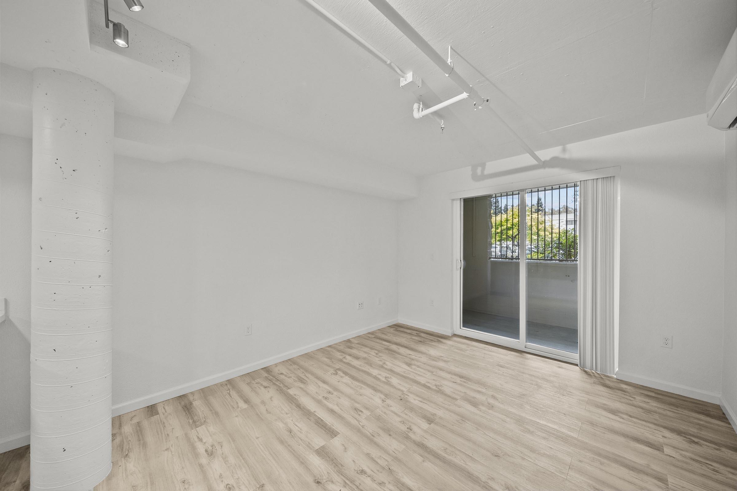A bright, empty interior room with light wooden flooring, white walls, and a concrete column. There are large sliding glass doors leading to a balcony, allowing natural light to fill the space. The overall ambiance is modern and minimalist.