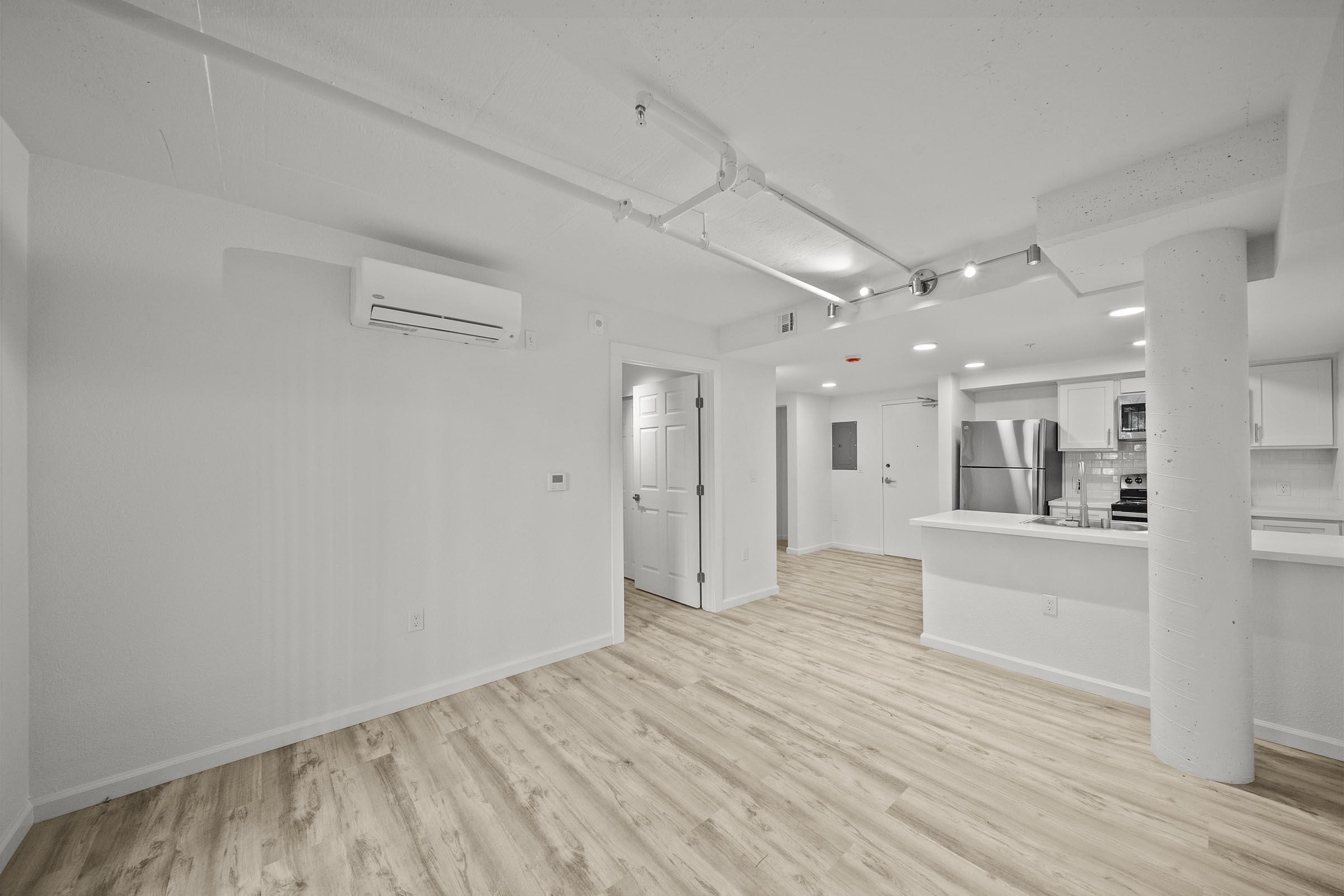 Modern empty room with light wood flooring, white walls, and a column. A doorway leads to a kitchen equipped with stainless steel appliances, visible in the background. Soft lighting fixtures are installed on the ceiling, and an air conditioning unit is mounted on the wall.
