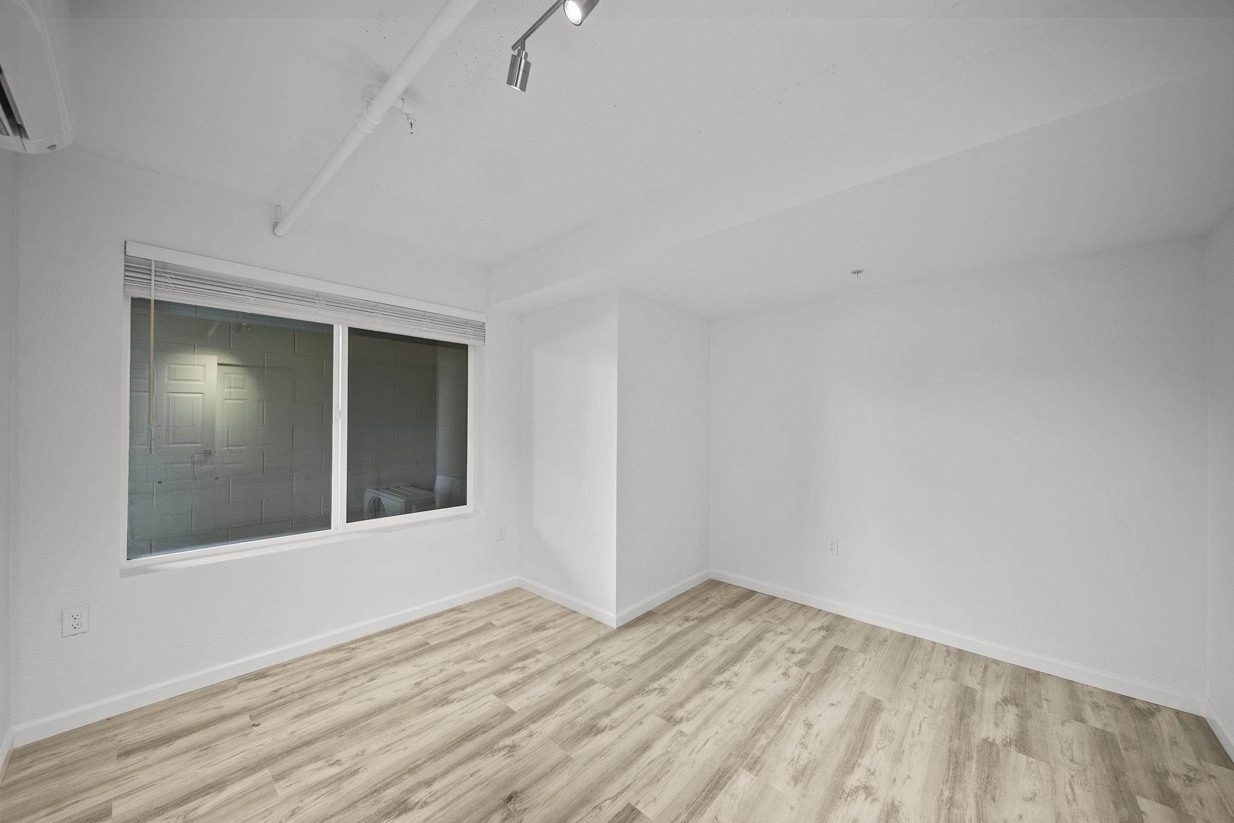 A blank, unfurnished room with light-colored walls and a wooden floor. There is a large window on one wall allowing natural light to enter, and a ceiling with exposed ductwork. The space appears modern and minimalistic, suitable for various uses or future decoration.