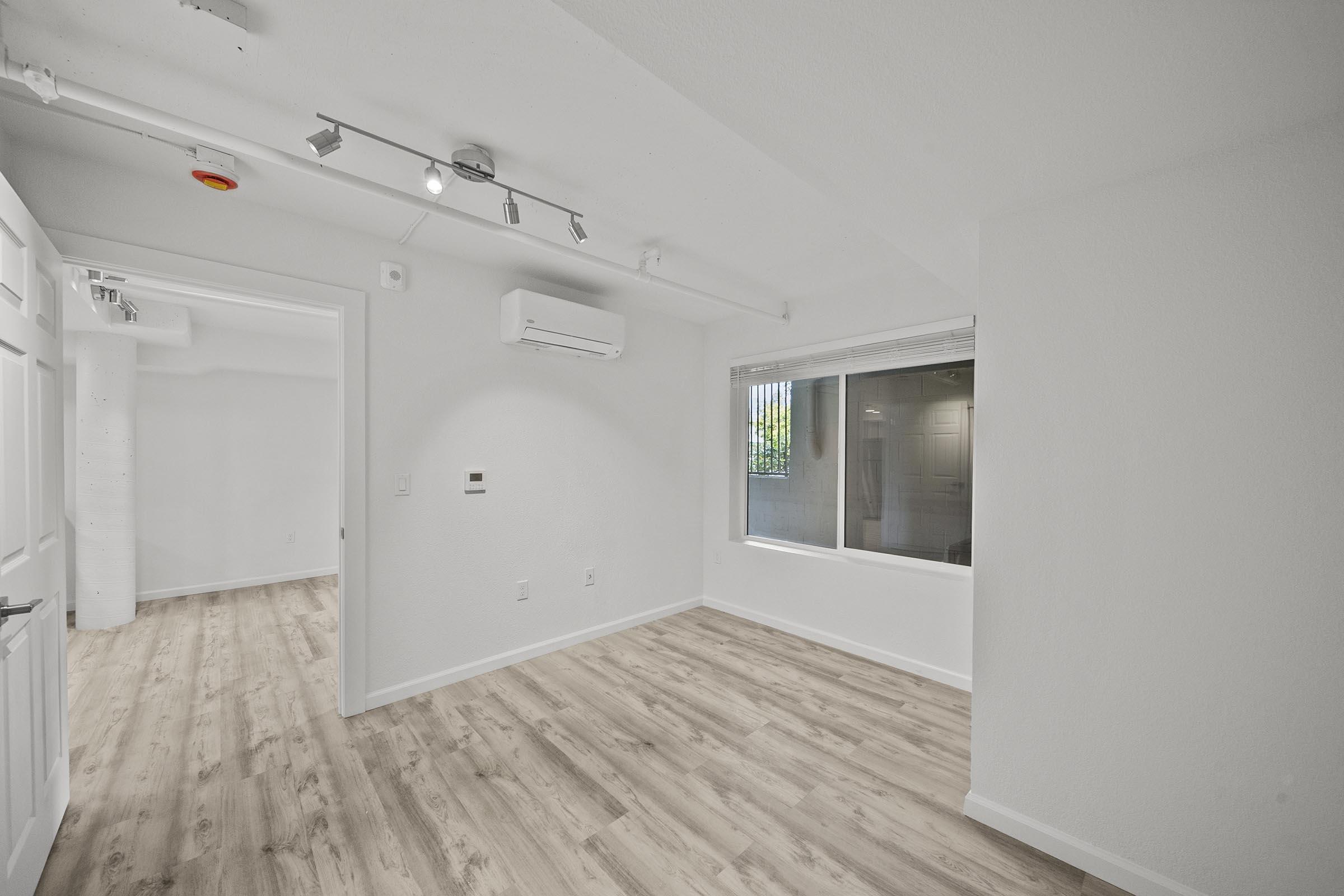 A bright, empty room with light-colored walls and wood-like flooring. There is a large window with blinds on one wall, providing natural light. An air conditioning unit is mounted near the ceiling. Another room is visible through an open door, indicating a spacious layout.