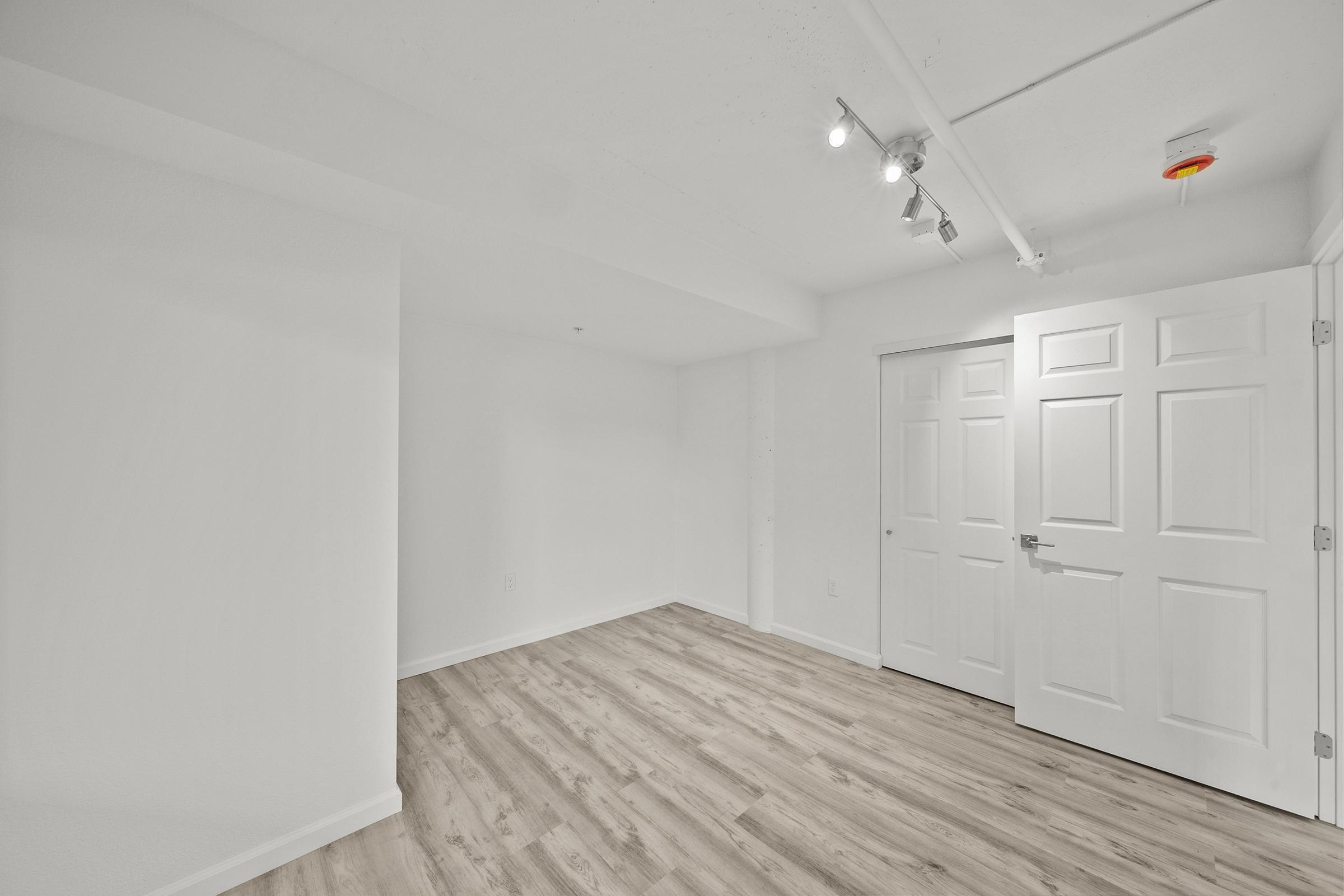 A spacious, empty room with white walls and a light wooden floor. Two doors are visible: one is a double door leading to a closet, while the other is a standard door. The ceiling features track lighting and there is a small overhead light. The room is well-lit and has a clean, modern aesthetic.