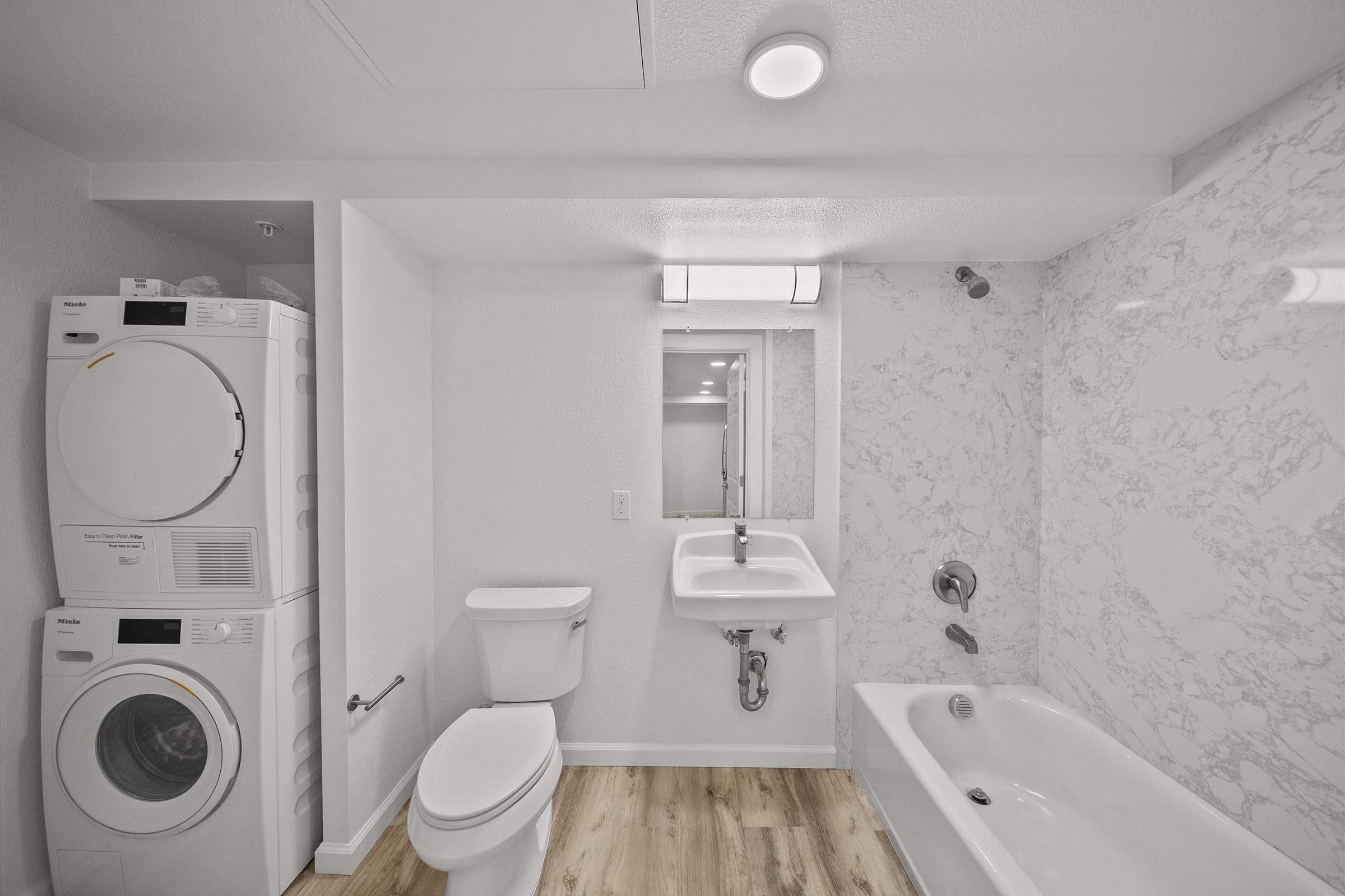 A modern bathroom featuring a white bathtub, a sink with a wall-mounted faucet, and a toilet. To the left, there is a stacked washing machine and dryer. The space has light marble walls, a large mirror, and wooden flooring, creating a clean and contemporary aesthetic.