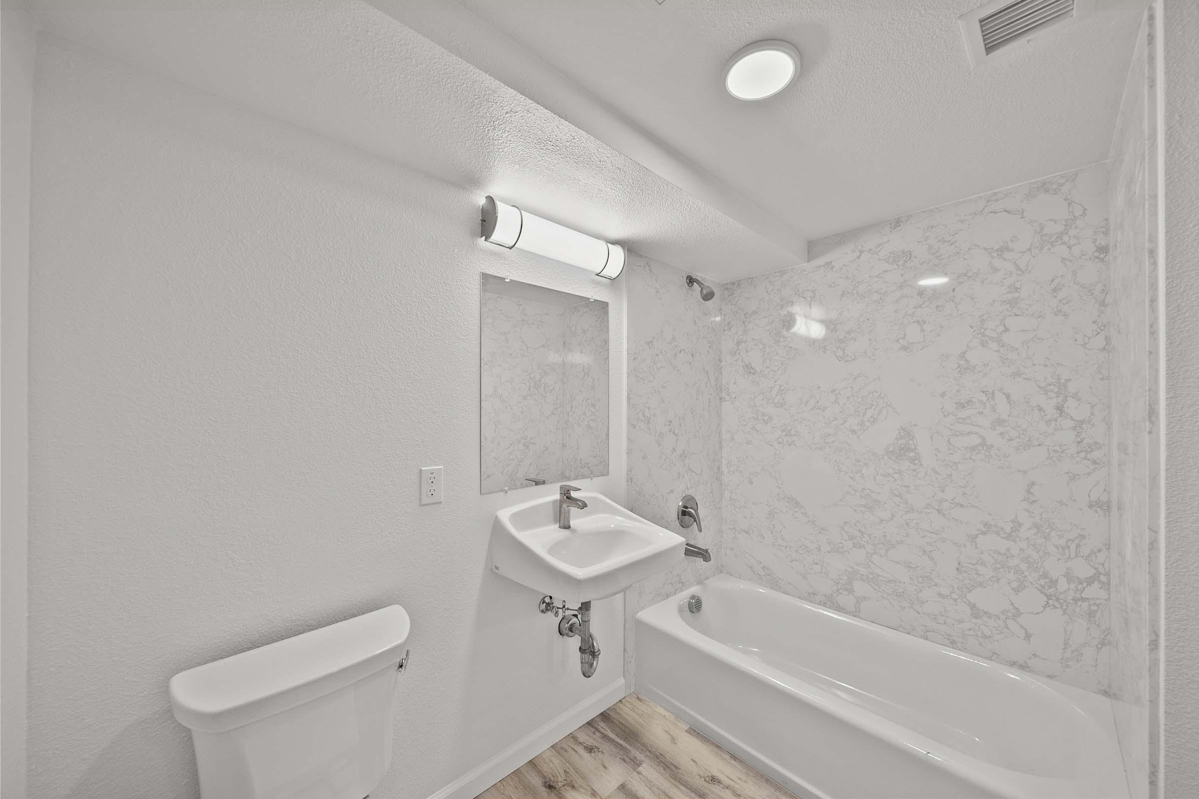 A modern bathroom featuring a white marble wall, a bathtub with a shower, a wall-mounted sink, a toilet, and overhead lighting. The space is well-lit and has neutral-colored walls and wood-style flooring.