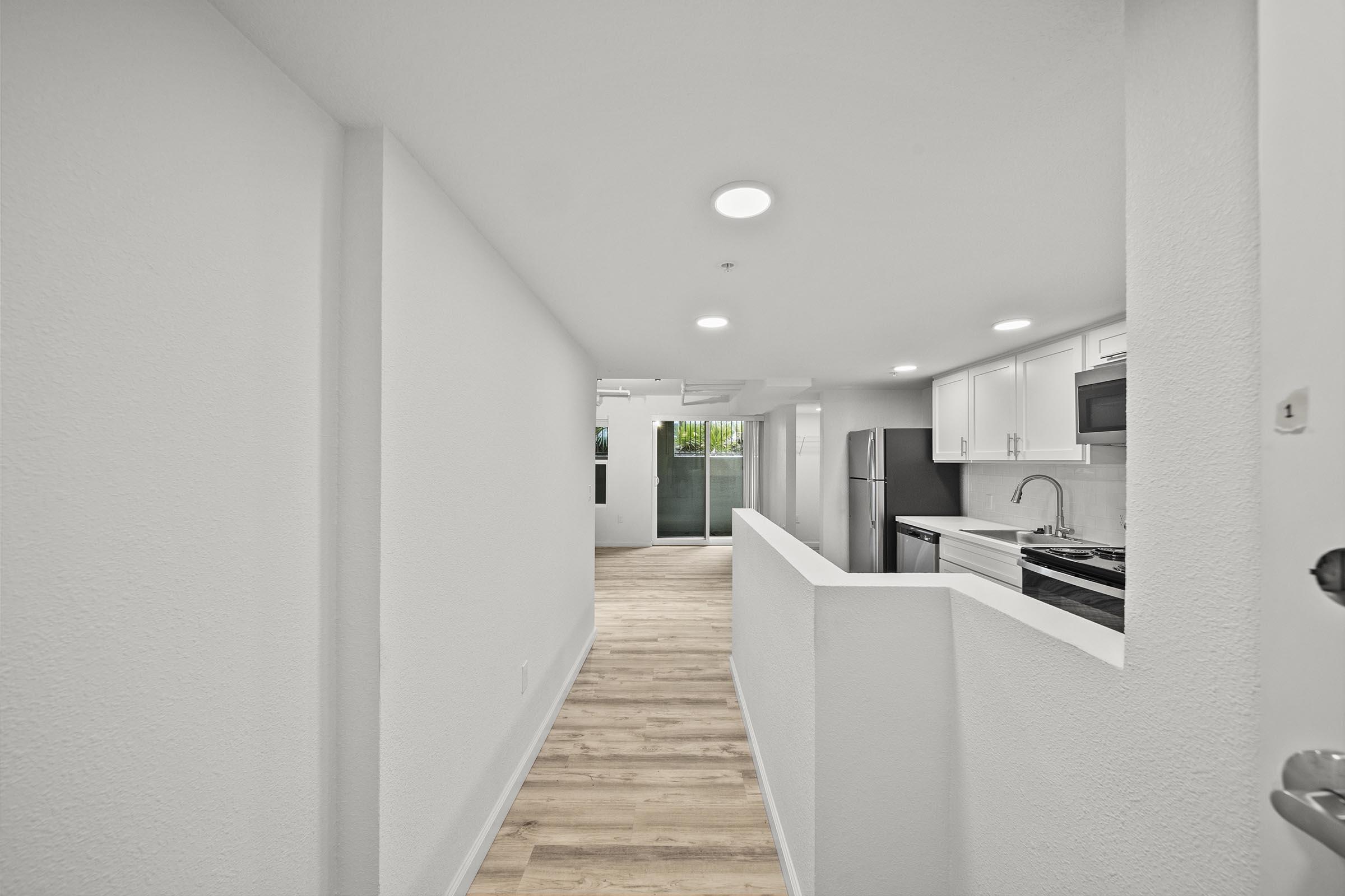 Interior view of a modern, bright hallway leading to an open-plan kitchen and living area. The walls are painted white, with recessed lighting and light wooden flooring. A large window at the end of the hallway allows natural light to fill the space, creating an inviting atmosphere.