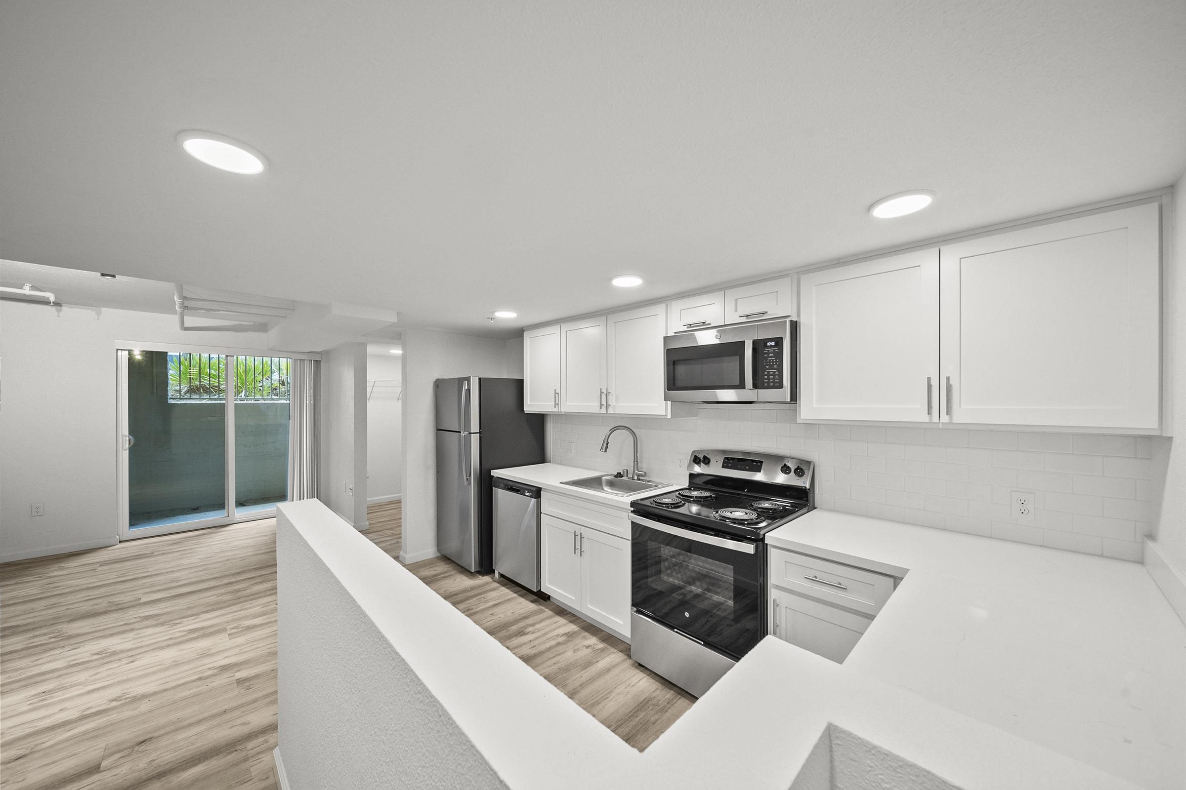 Modern kitchen featuring white cabinets, stainless steel appliances, a gas stove, and a large sink. The space includes a refrigerator and is open to a living area with light-colored wood flooring. Large windows in the background provide natural light, leading to a balcony.