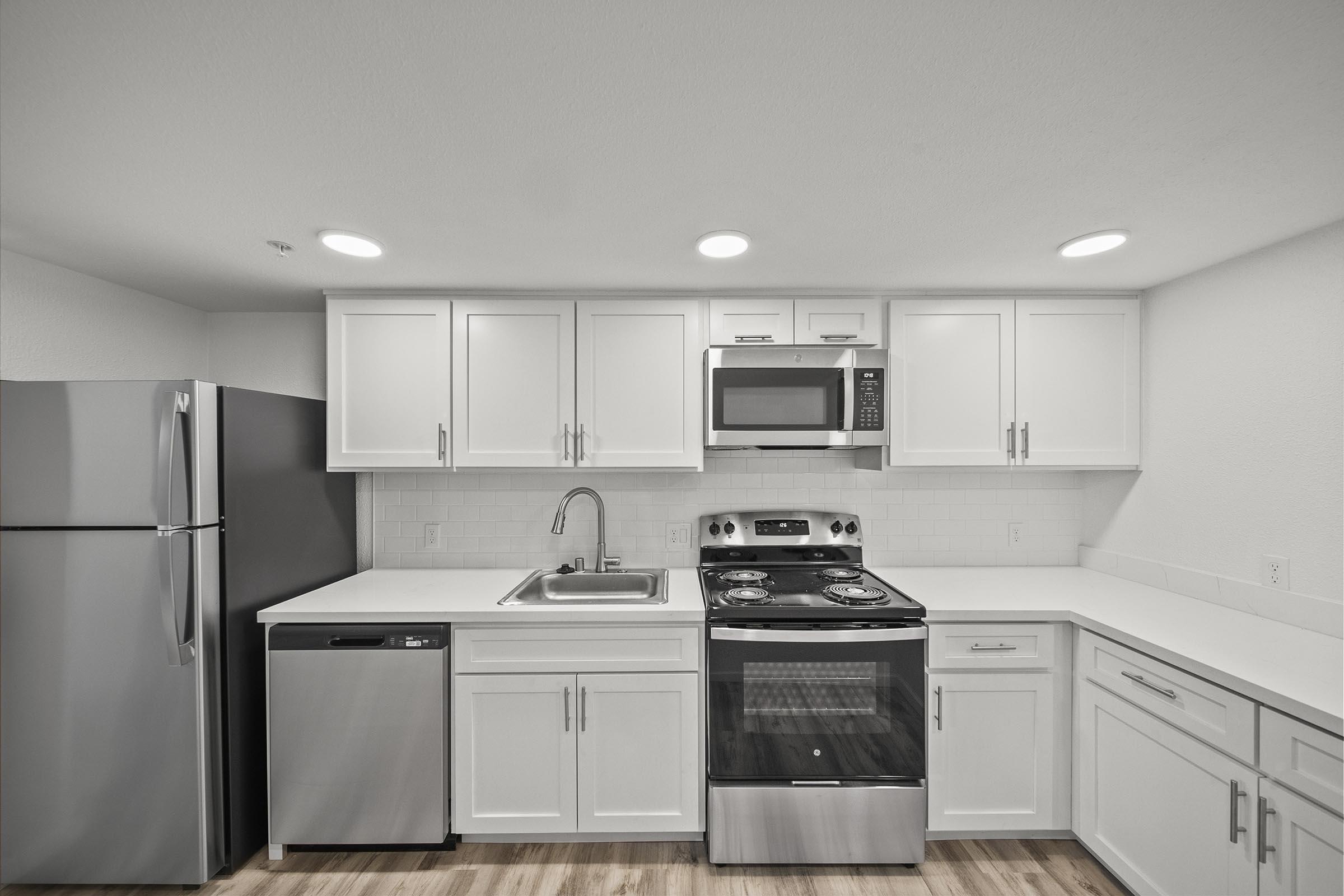 Modern kitchen featuring white cabinetry, a stainless steel refrigerator, a silver dishwasher, and a gas stove with an oven. The kitchen also includes a microwave and a stainless steel sink on a white countertop, illuminated by recessed lighting. Wood laminate flooring completes the contemporary look.