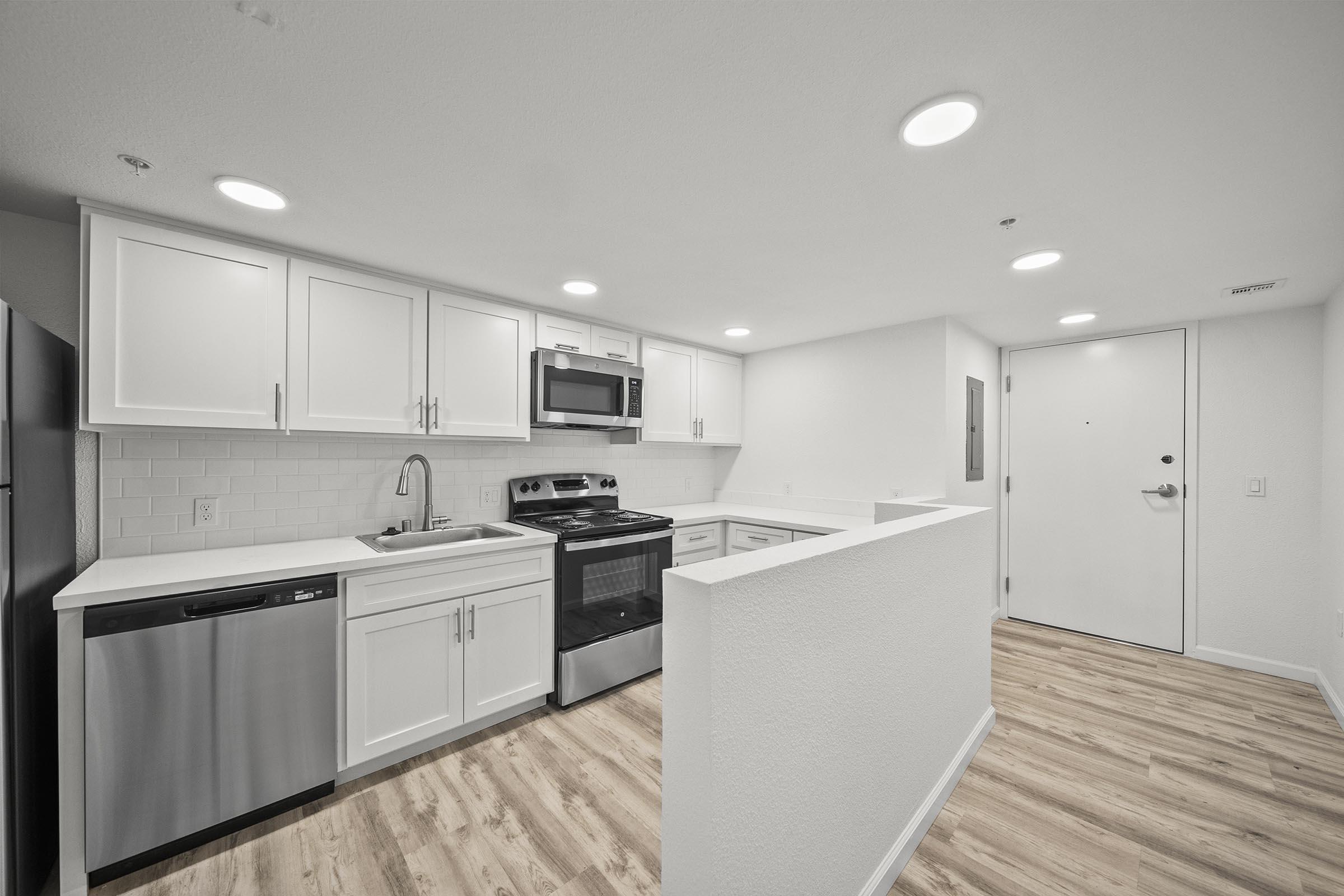 Modern kitchen featuring white cabinetry, stainless steel appliances including a refrigerator, stove, and dishwasher. The countertop is light-colored with a clean, minimalist design. The flooring is wood-like laminate, and overhead lights provide ample illumination. A door leads to the entryway.