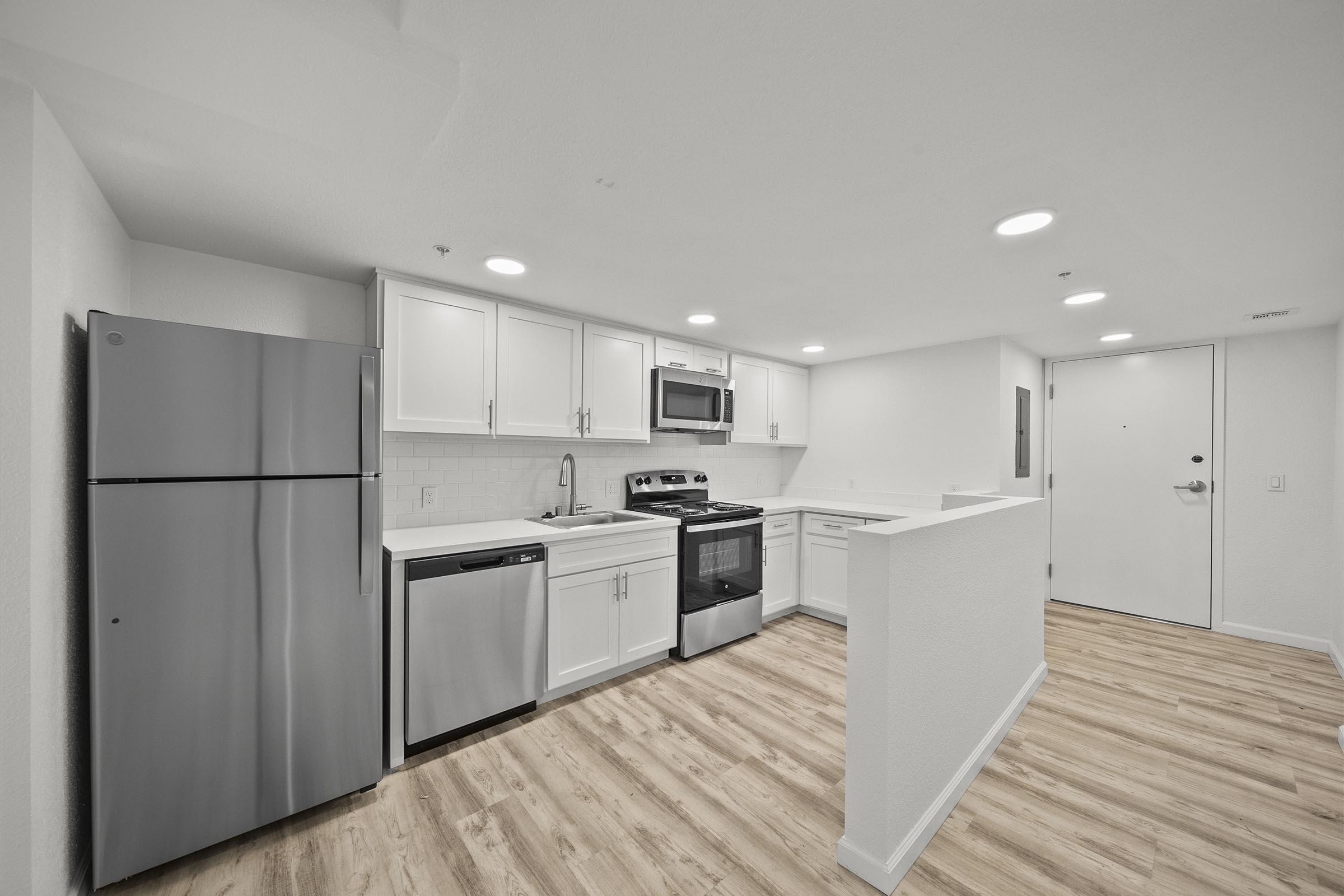 Modern kitchen featuring stainless steel appliances including a refrigerator, dishwasher, microwave, and oven. The space has light-colored cabinets and a spacious countertop with a wood-like flooring. Bright overhead lights illuminate the room, creating a welcoming atmosphere.