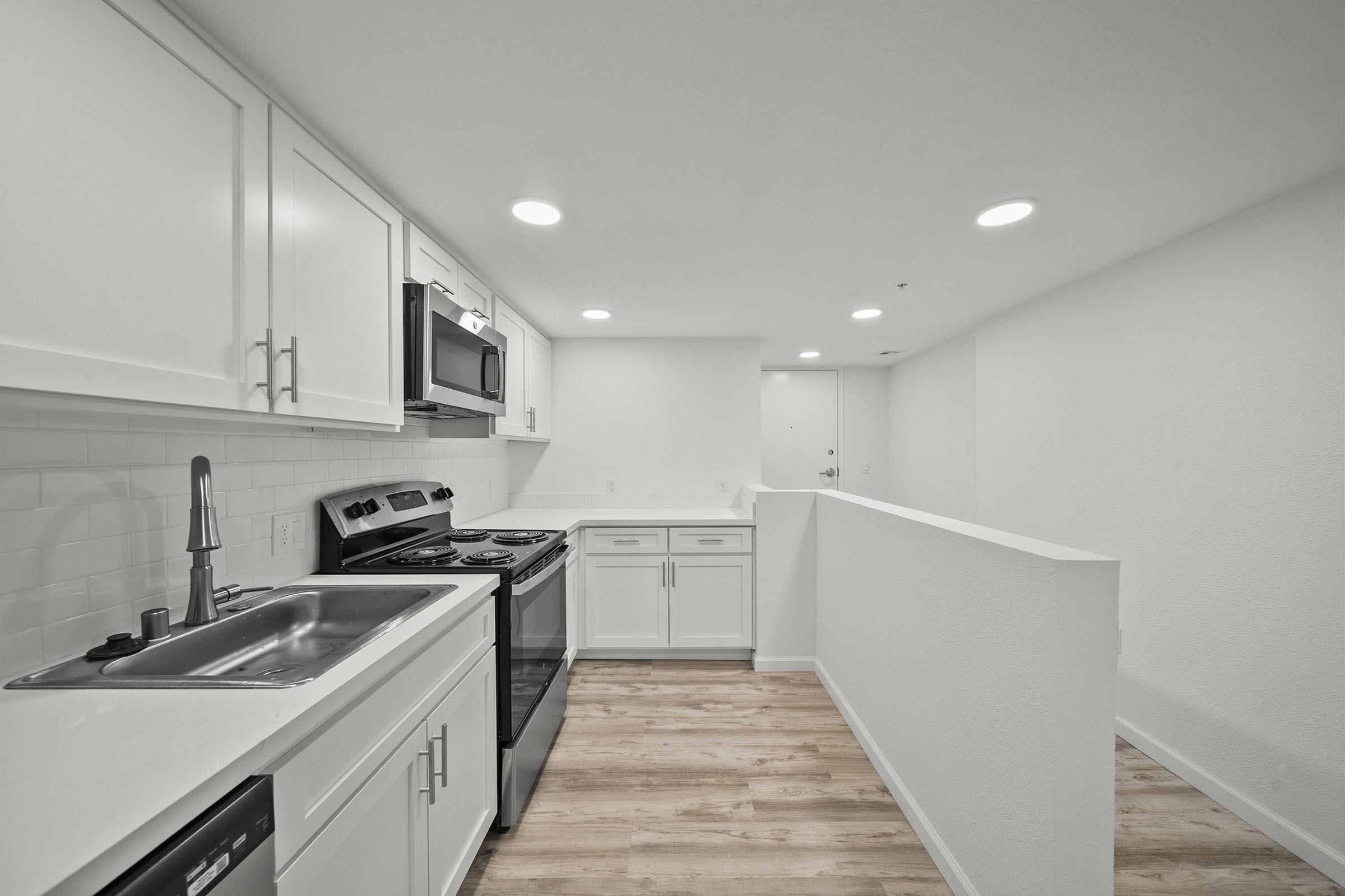 Modern kitchen with white cabinetry, stainless steel appliances, and a single basin sink. The layout features a breakfast bar and a light, airy atmosphere with neutral-colored walls and wood-like flooring. Bright recessed lighting enhances the spacious feel of the area.