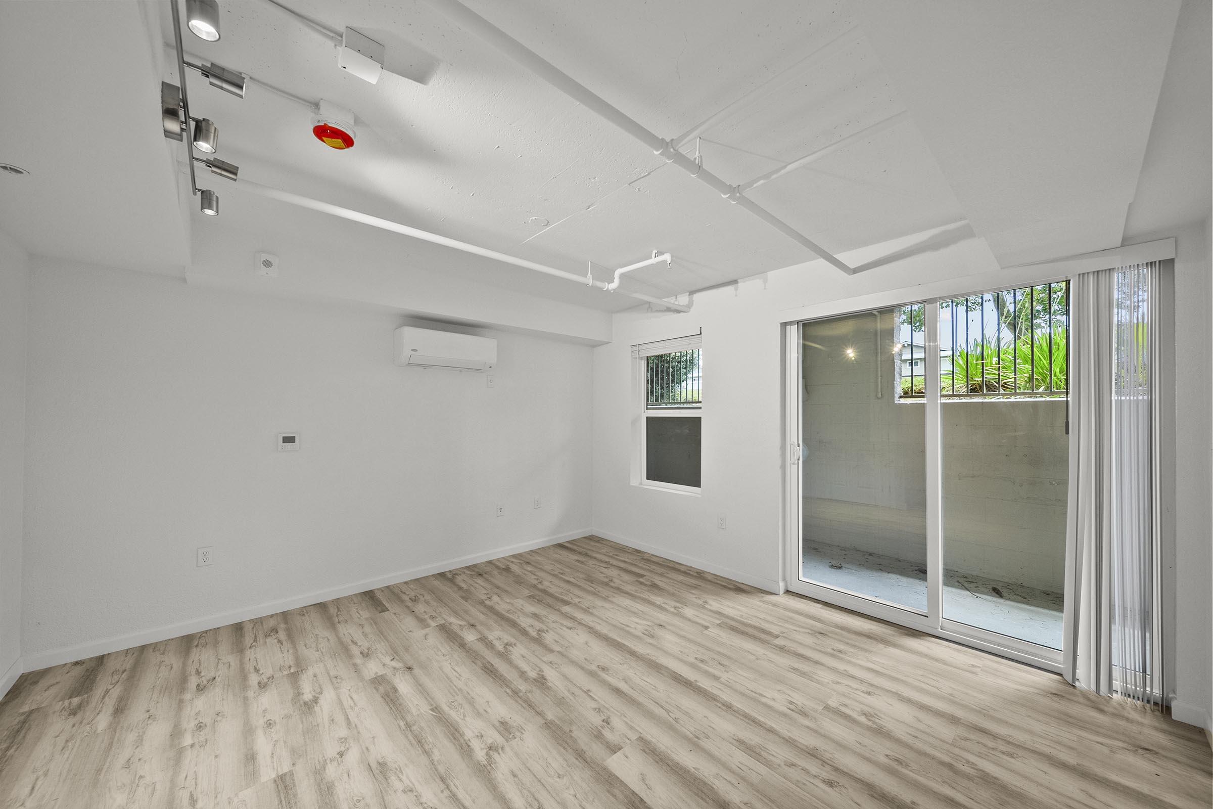 A spacious, empty room featuring light-colored walls and wooden flooring. There is an air conditioning unit on the wall, track lighting on the ceiling, and large glass sliding doors that lead to a small outdoor area. The room is bright with natural light flowing in.