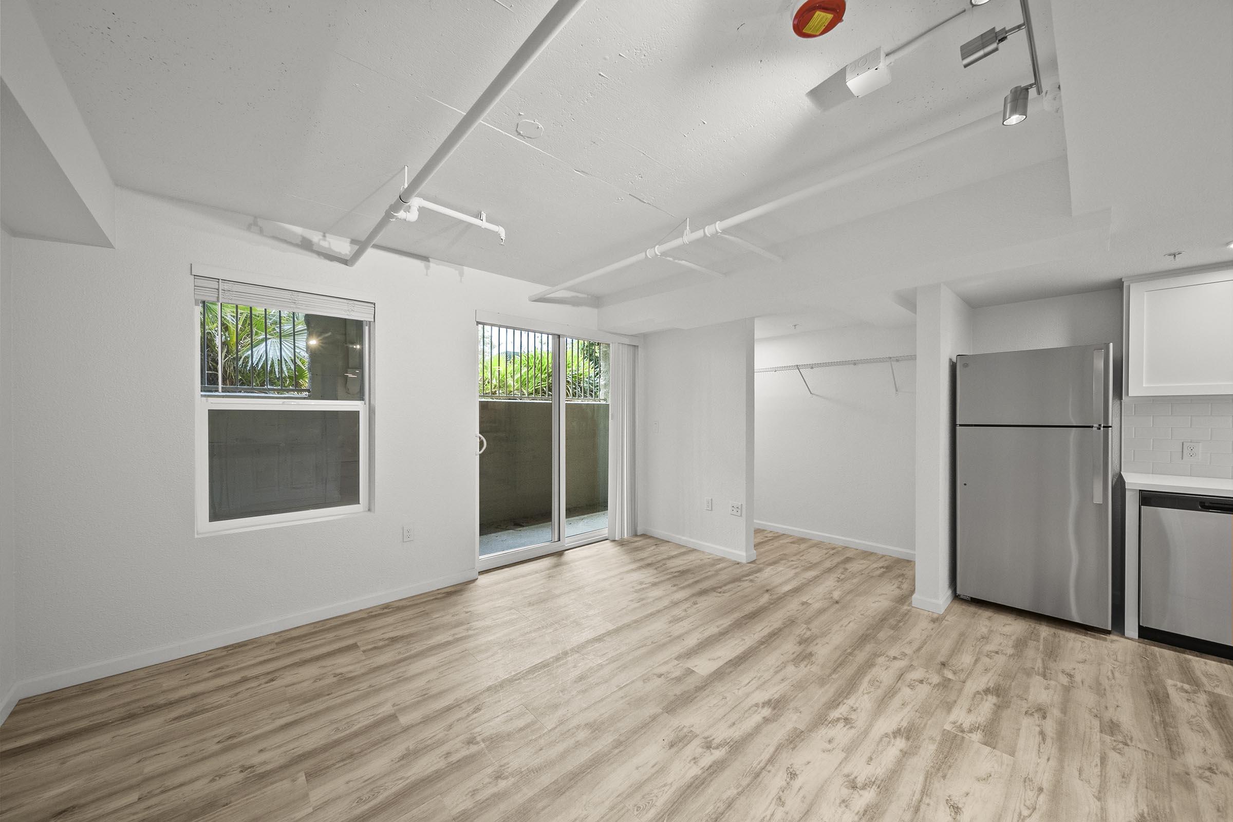 A spacious, empty room with light wood flooring, white walls, and large windows. It features a sliding glass door leading to an outdoor area, a modern stainless steel refrigerator, and overhead lighting. The layout is open, creating a bright and inviting atmosphere.