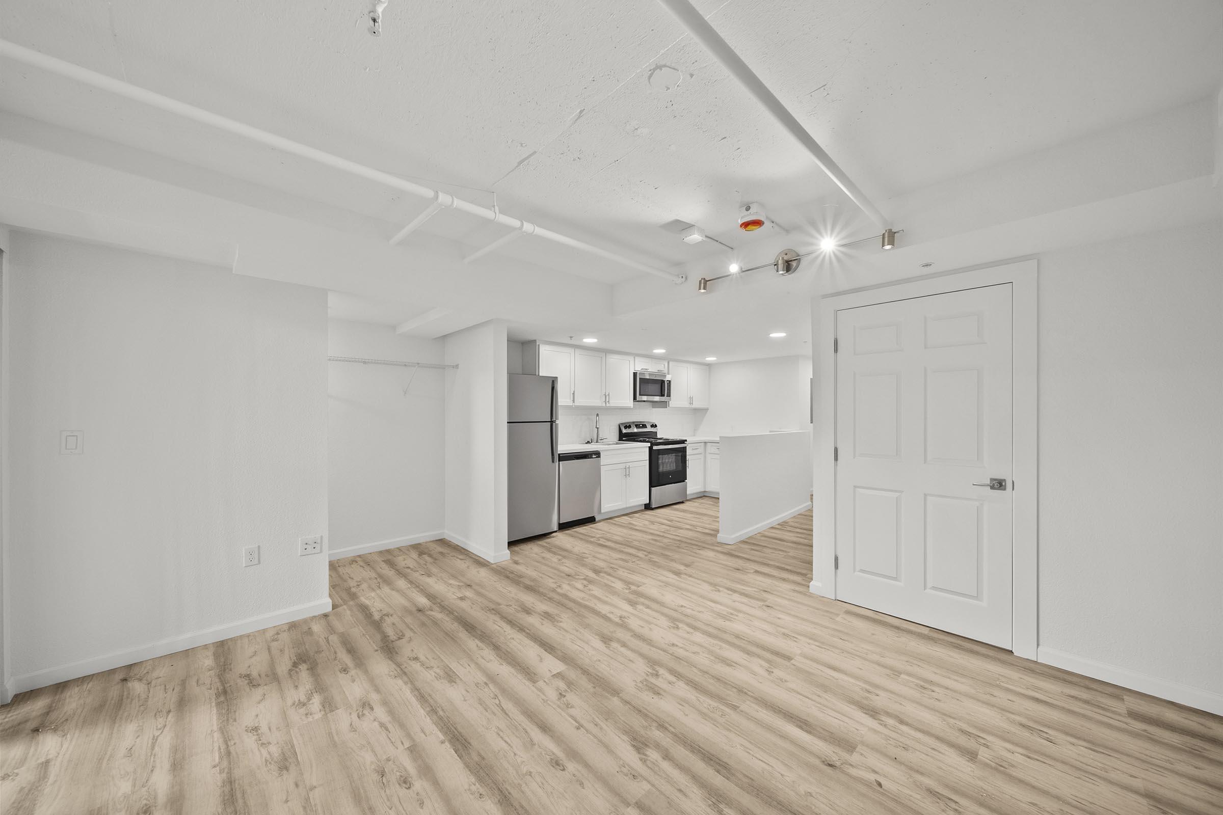 A spacious, modern basement apartment featuring light hardwood flooring, a kitchen with stainless steel appliances, white cabinetry, and bright lighting. The open layout includes a living area and a door leading to another room or closet space. The walls are painted in a crisp, clean white.