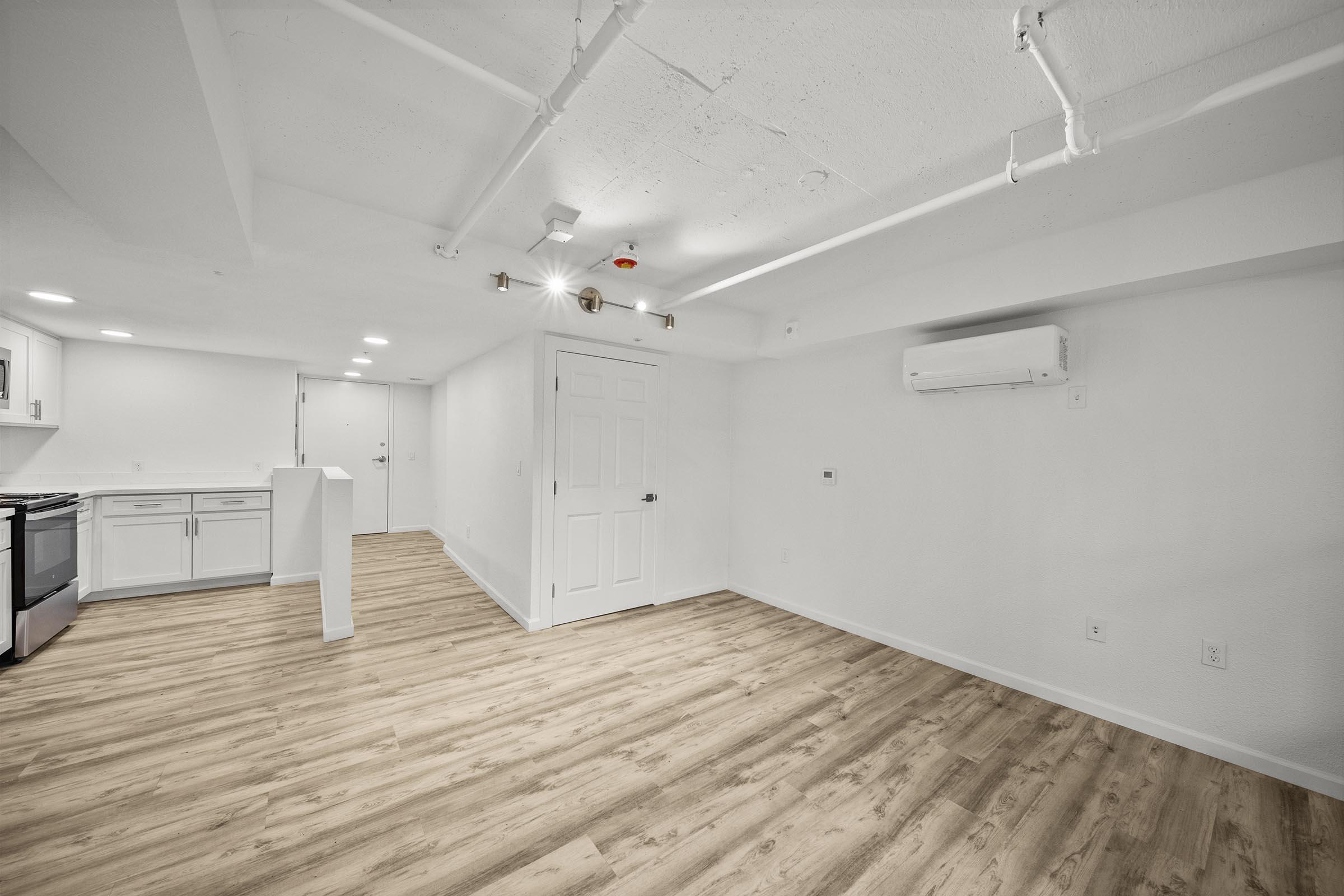 Bright, modern interior of an open-concept space featuring light-colored walls, laminate flooring, and recessed lighting. The kitchen area includes a countertop and appliances. There is an air conditioning unit on the wall, and the space appears freshly renovated and uncluttered, ideal for various uses.