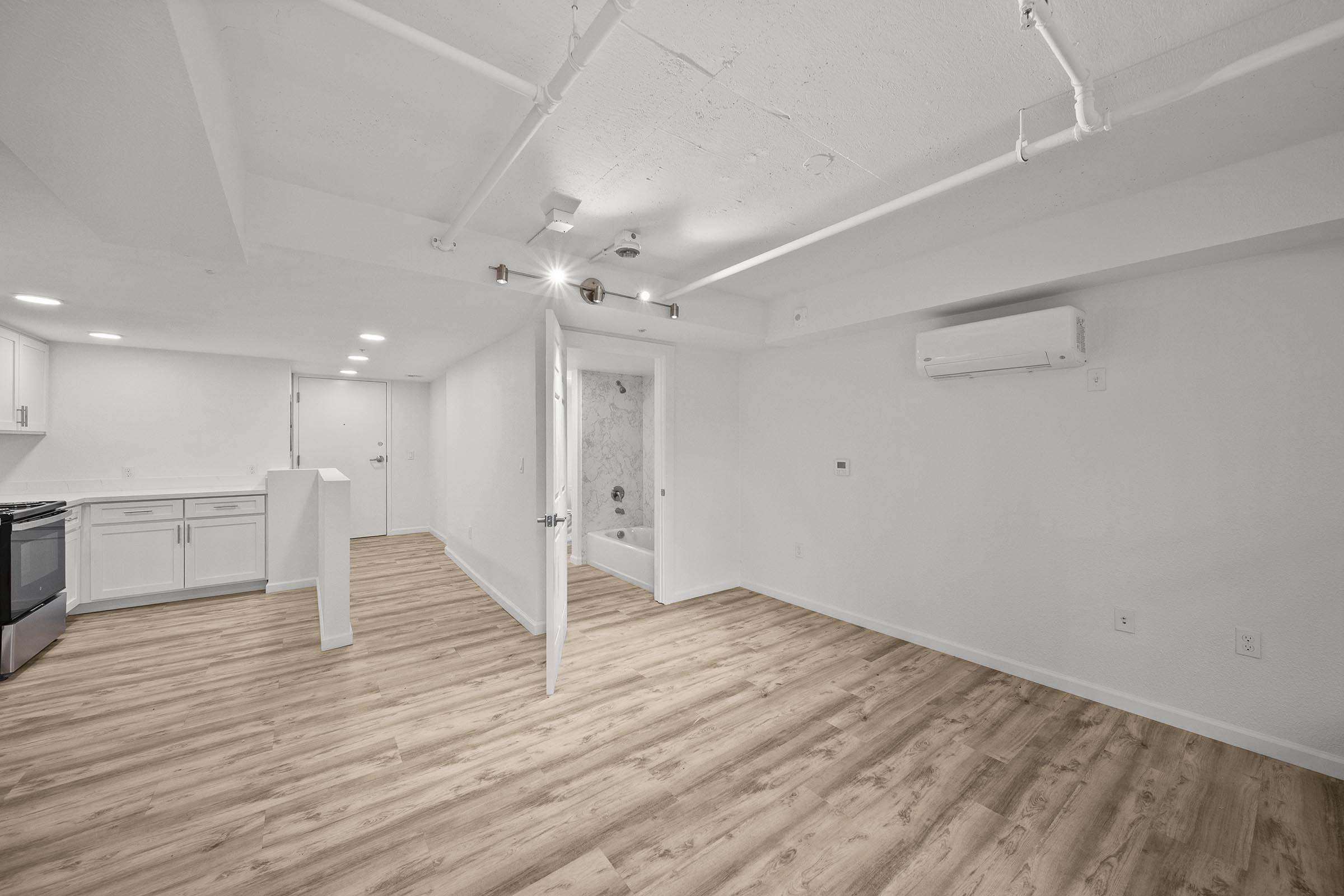 A spacious, modern apartment interior featuring light-colored walls and wood-style flooring. The open layout includes a kitchen area with white cabinets and appliances, a bright living space, and a bathroom visible through an open door. Natural light fills the room, creating an inviting atmosphere.