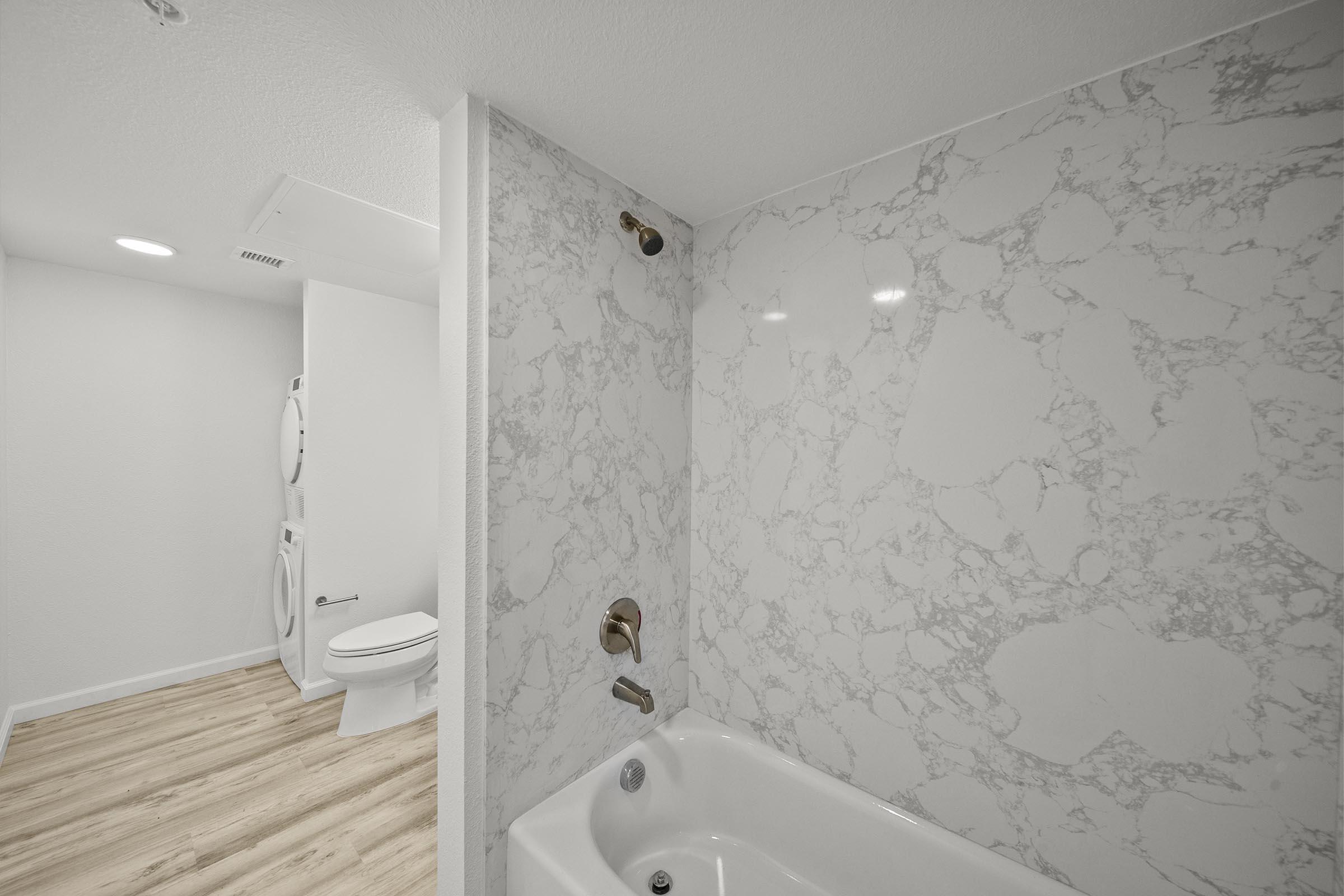 A modern bathroom featuring a bathtub with a marble-patterned wall, a gold shower fixture, and wooden flooring. In the background, a toilet is visible alongside a stackable washer and dryer unit, all in a bright, well-lit space.
