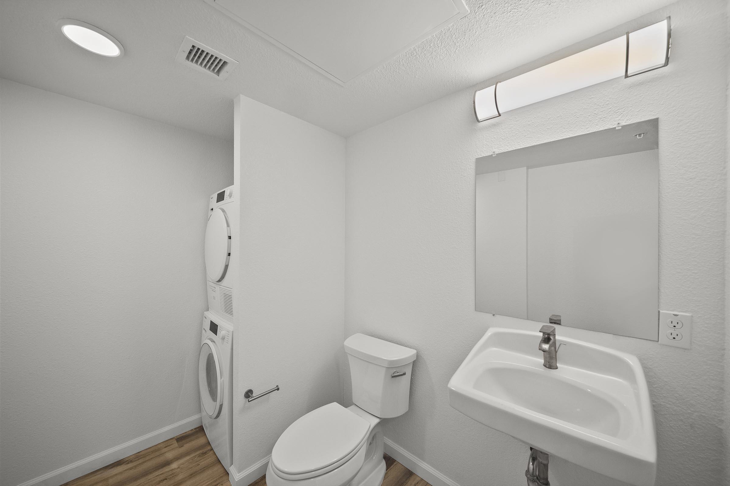 A clean and modern bathroom featuring a toilet, a small sink with a faucet, a mirror, and a stacked washer and dryer unit. The walls are painted white, and the flooring is light wood. A ceiling light fixture provides illumination in the room.