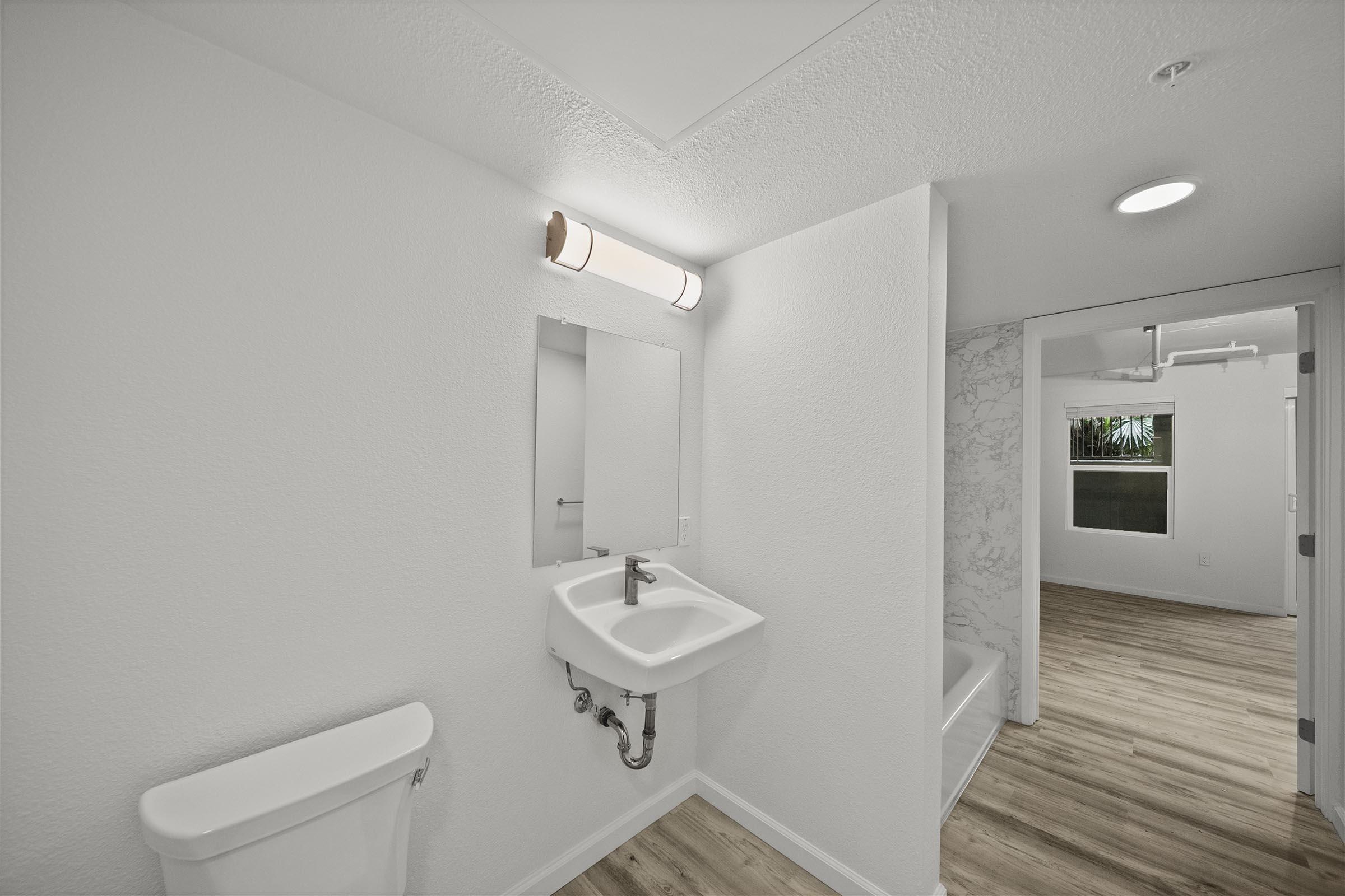 A clean, modern bathroom featuring a white sink with a mirror above it, a white toilet, and light wood flooring. The walls are painted white, and there is a marble accent wall visible. A doorway leads to another room with a window, providing natural light.