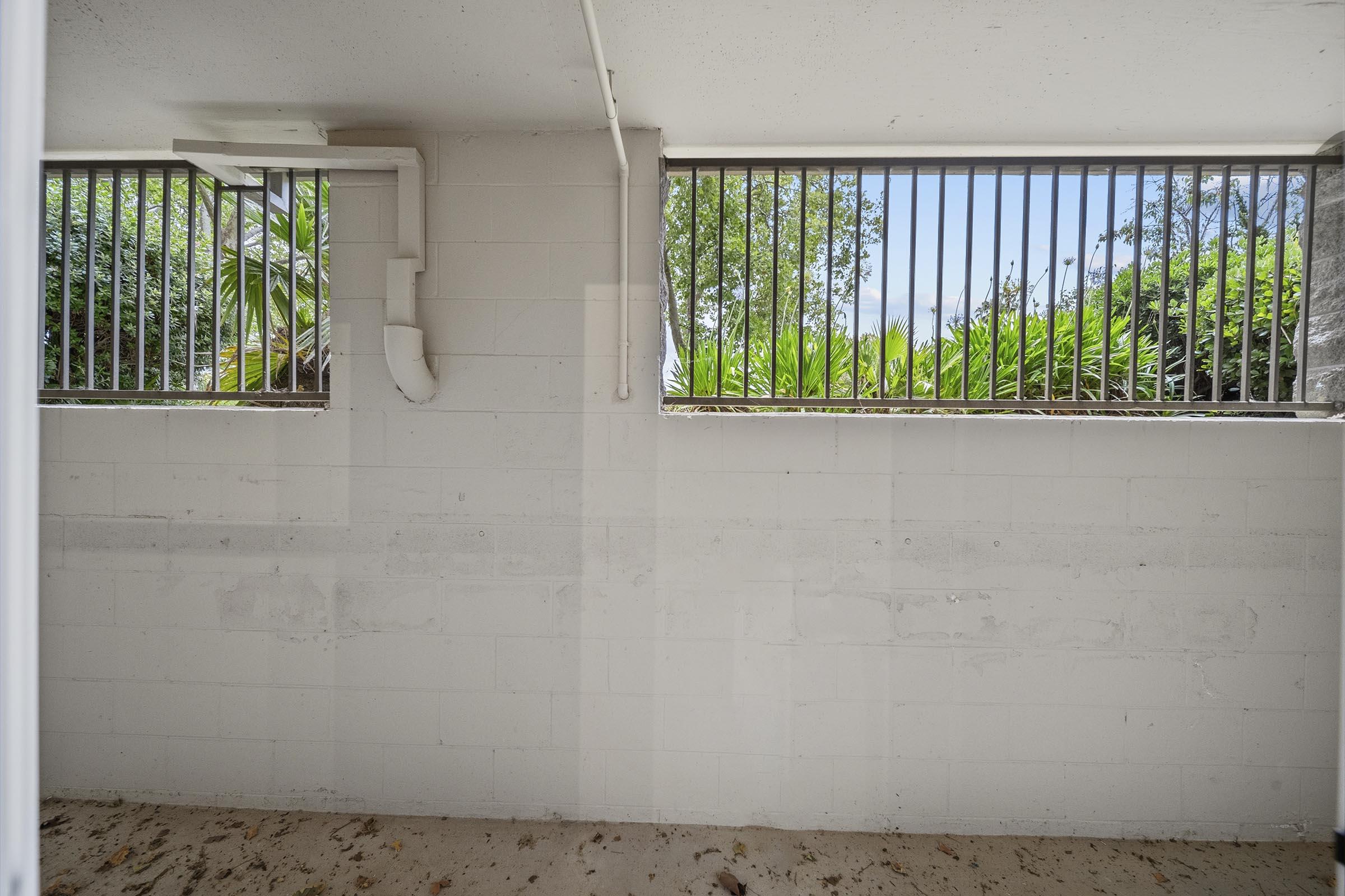 An empty interior space with two barred windows allowing light in. The walls are painted white with visible concrete blocks. Outside the windows, green foliage can be seen. The floor is bare, indicating a lack of furnishings or decoration.