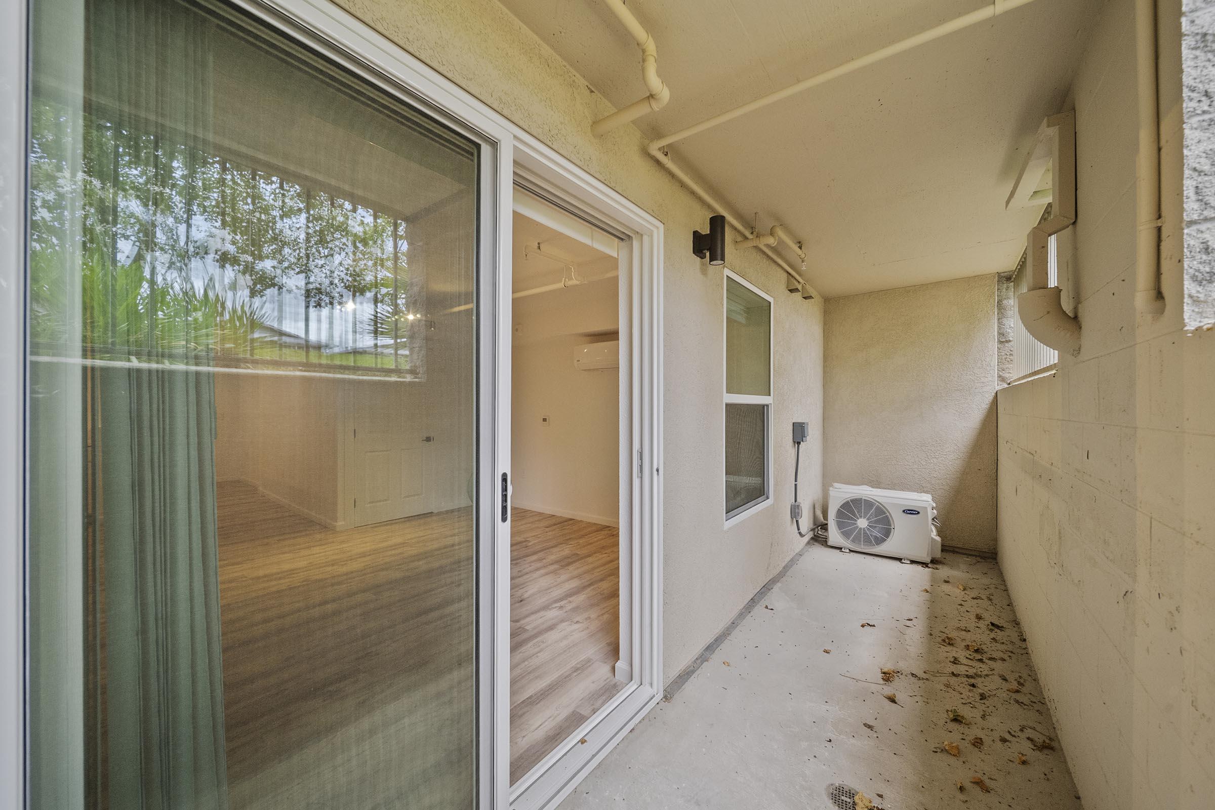 View of a small balcony or patio area with sliding glass doors leading to an interior room. The space features a light-colored wall, an air conditioning unit, and a floor with scattered leaves. Natural light filters through the doors, reflecting on the interior wooden flooring.