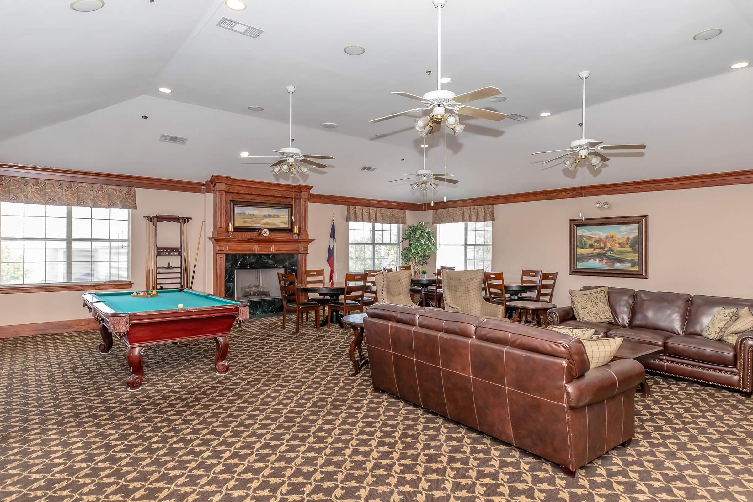 A spacious common area featuring a billiards table, a comfortable leather couch, and wooden dining tables. Large windows allow natural light to fill the room, while ceiling fans provide airflow. The decor includes paintings and potted plants, creating a warm and inviting atmosphere.