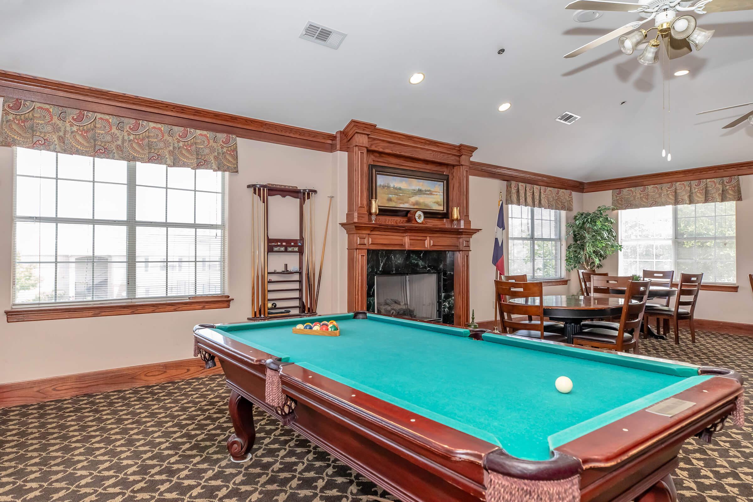 A cozy recreational room featuring a green felt pool table with balls set up, a wooden chair set nearby, and a fireplace decorated with a painting. Large windows with floral curtains let in natural light, and a plant decorates the corner, adding to the warm ambiance.