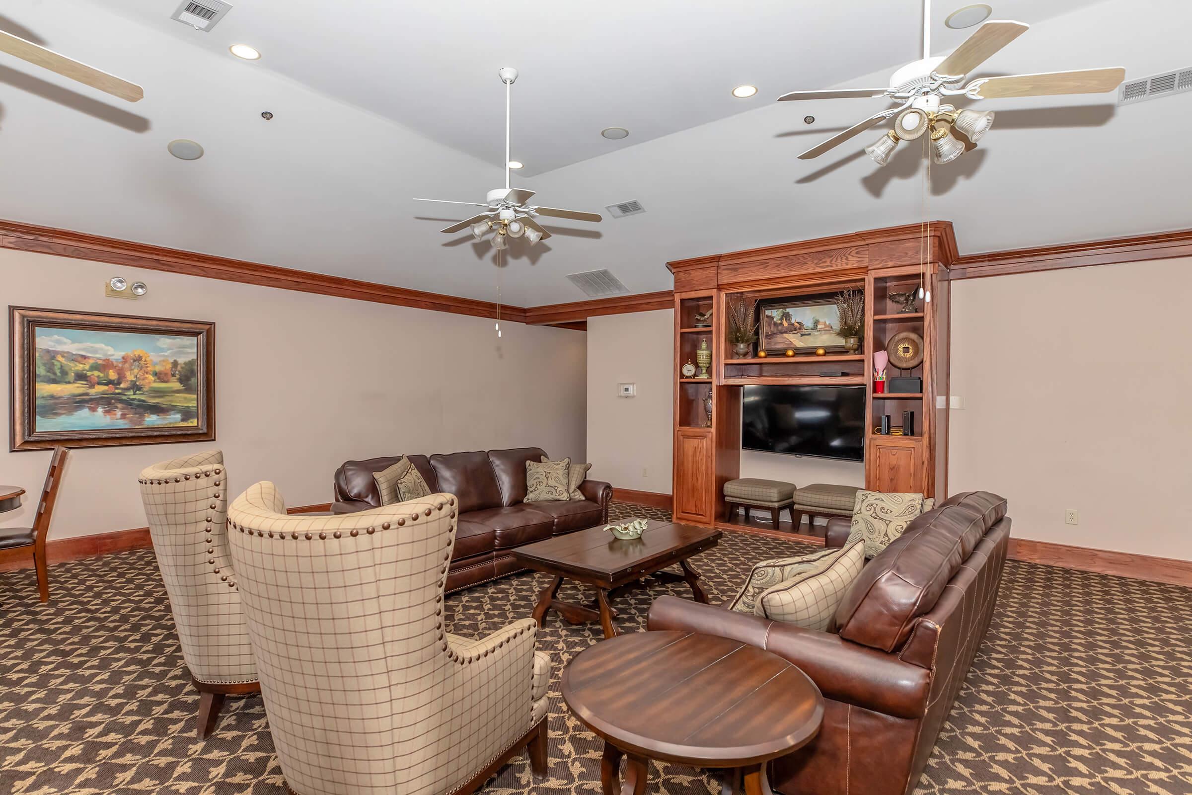 A cozy living room featuring a mix of leather and upholstered furniture, a wooden entertainment unit, and decorative art. The space is well-lit with ceiling fans, has a neutral color palette, and a patterned carpet, creating a welcoming atmosphere for relaxation and conversation.