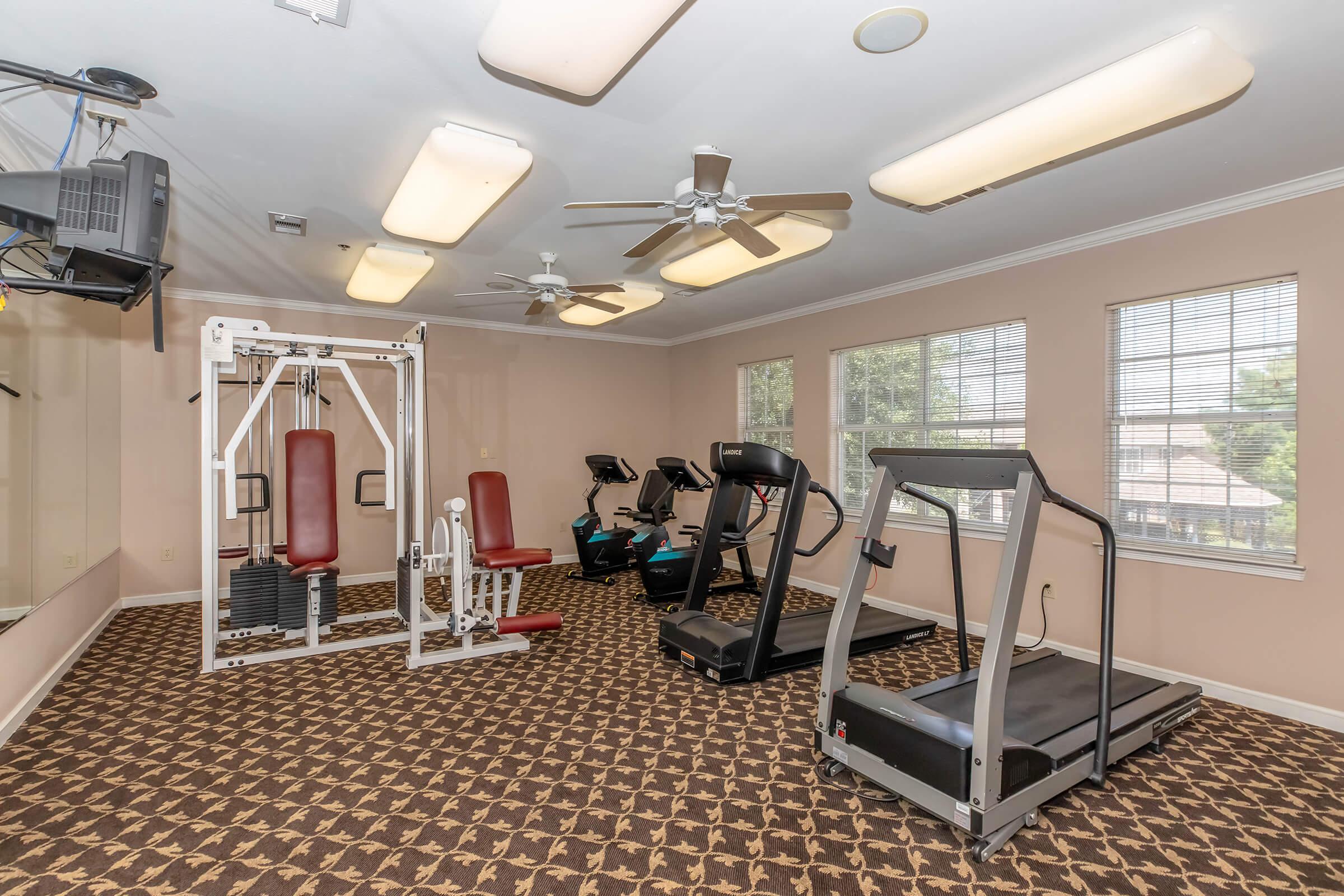 A fitness room featuring exercise equipment, including a weight machine, treadmills, and stationary bikes. The space has large windows for natural light, ceiling fans, and a patterned carpet, creating a bright and inviting atmosphere for workouts.