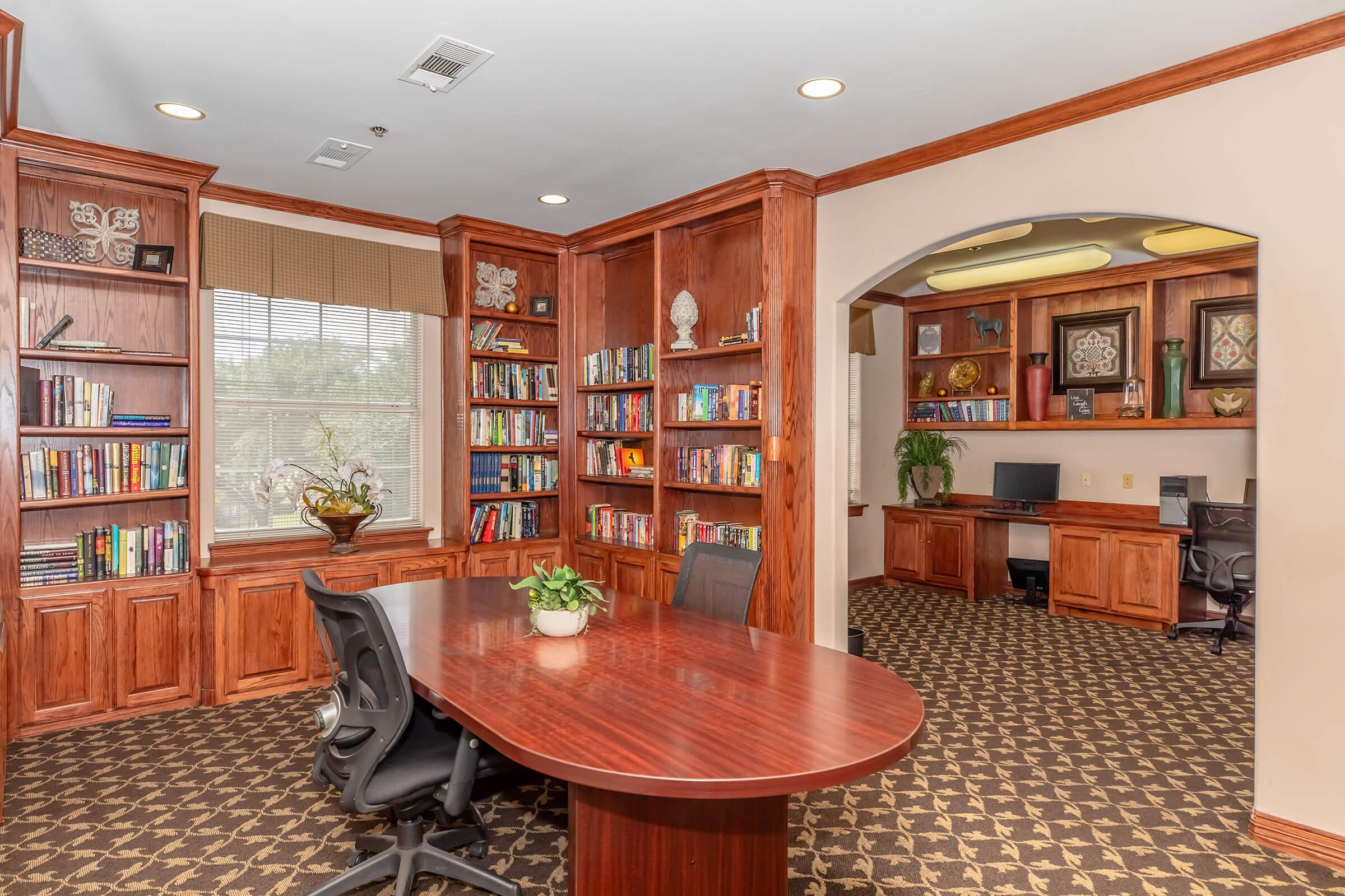 A cozy office space featuring wooden shelves filled with books, a large round table with black chairs, a potted plant in the center, and a window bringing in natural light. An adjacent area includes a desk against the wall, enhancing the organized and inviting atmosphere of the workspace.