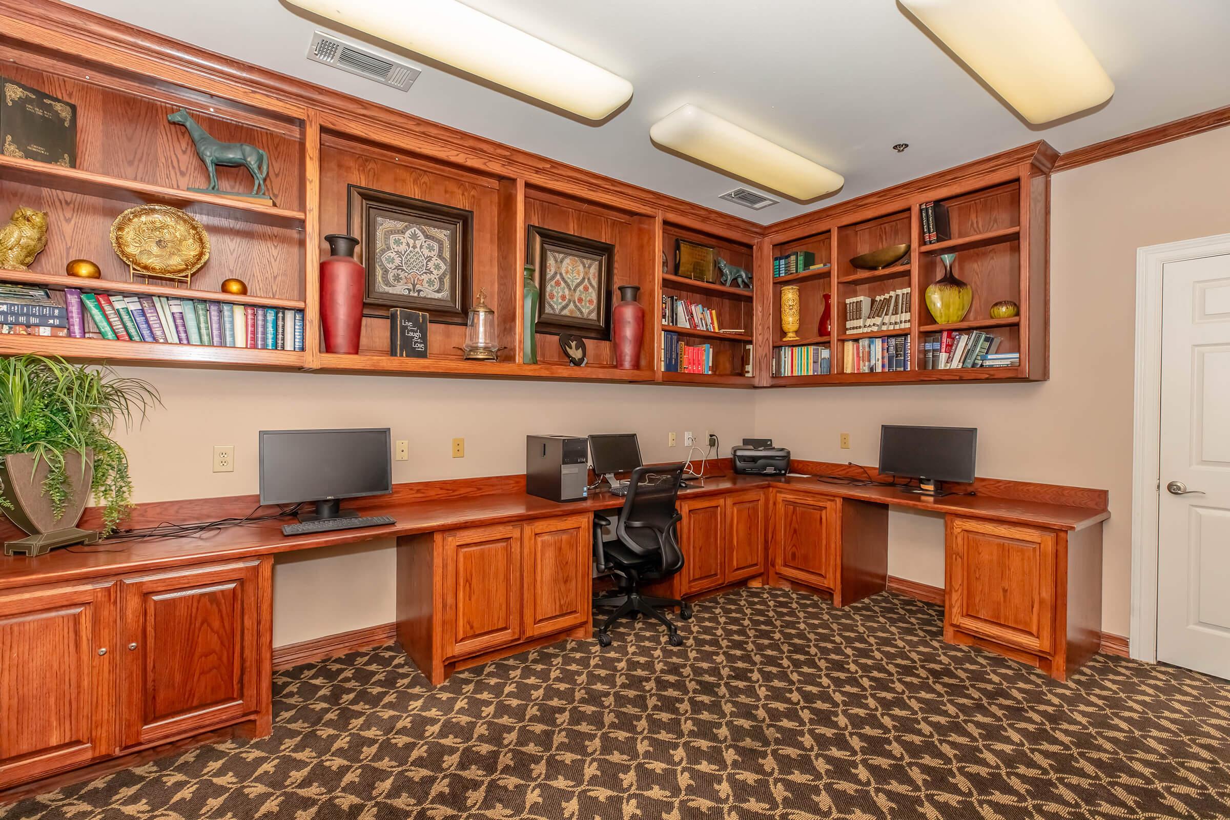 A spacious home office featuring wooden cabinetry and shelves filled with decorative items and books. Two computer stations with monitors are positioned on the wooden desks. The room has a patterned carpet and warm lighting, creating a cozy yet functional workspace.
