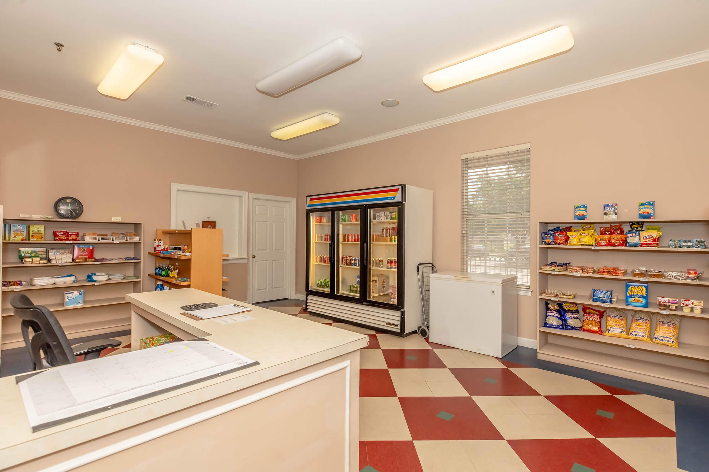 Interior of a convenience store or pantry area featuring shelves stocked with snacks and drinks. A refrigerator with colorful doors is visible, holding beverages. The layout includes a front counter with a phone and a large freezer nearby. The overall color scheme is light with patterned flooring.