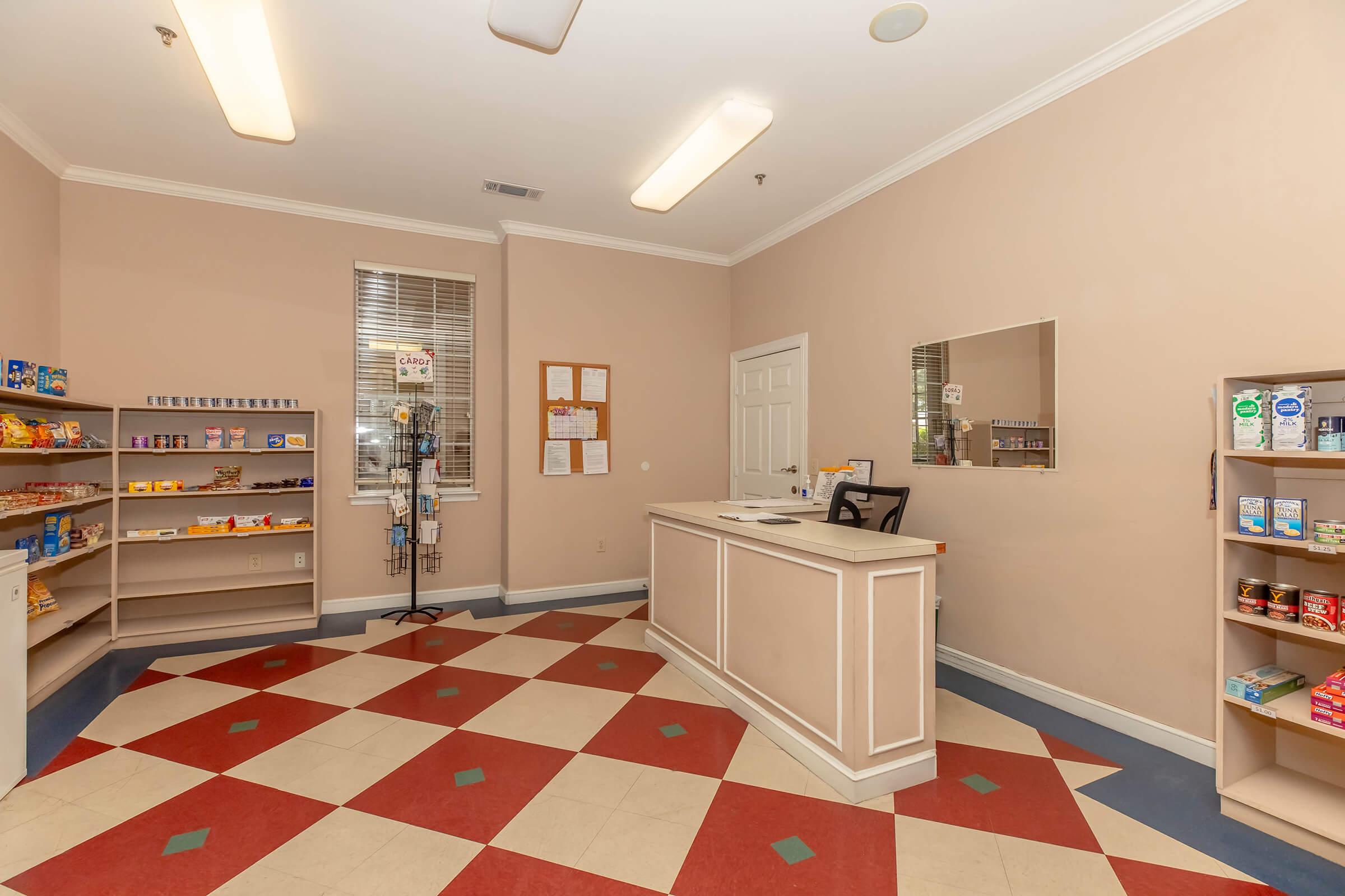Interior view of a small convenience store or shop, featuring a reception counter, food items displayed on shelves, and a tiled floor with a red and cream checkerboard pattern. The walls are painted a soft beige, and there is a mirror on the back wall along with some notice boards.