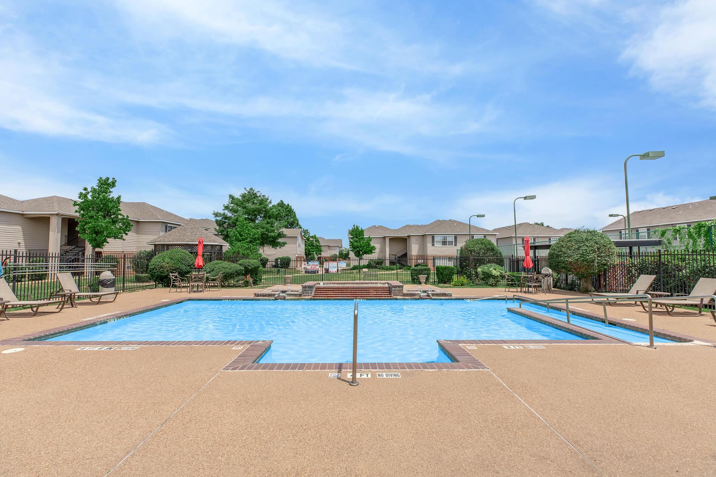 A bright outdoor swimming pool surrounded by lounge chairs and elegant landscaping. The pool area features pathways, trees, and residential buildings in the background under a clear blue sky. Red umbrellas provide shaded seating near the pool.