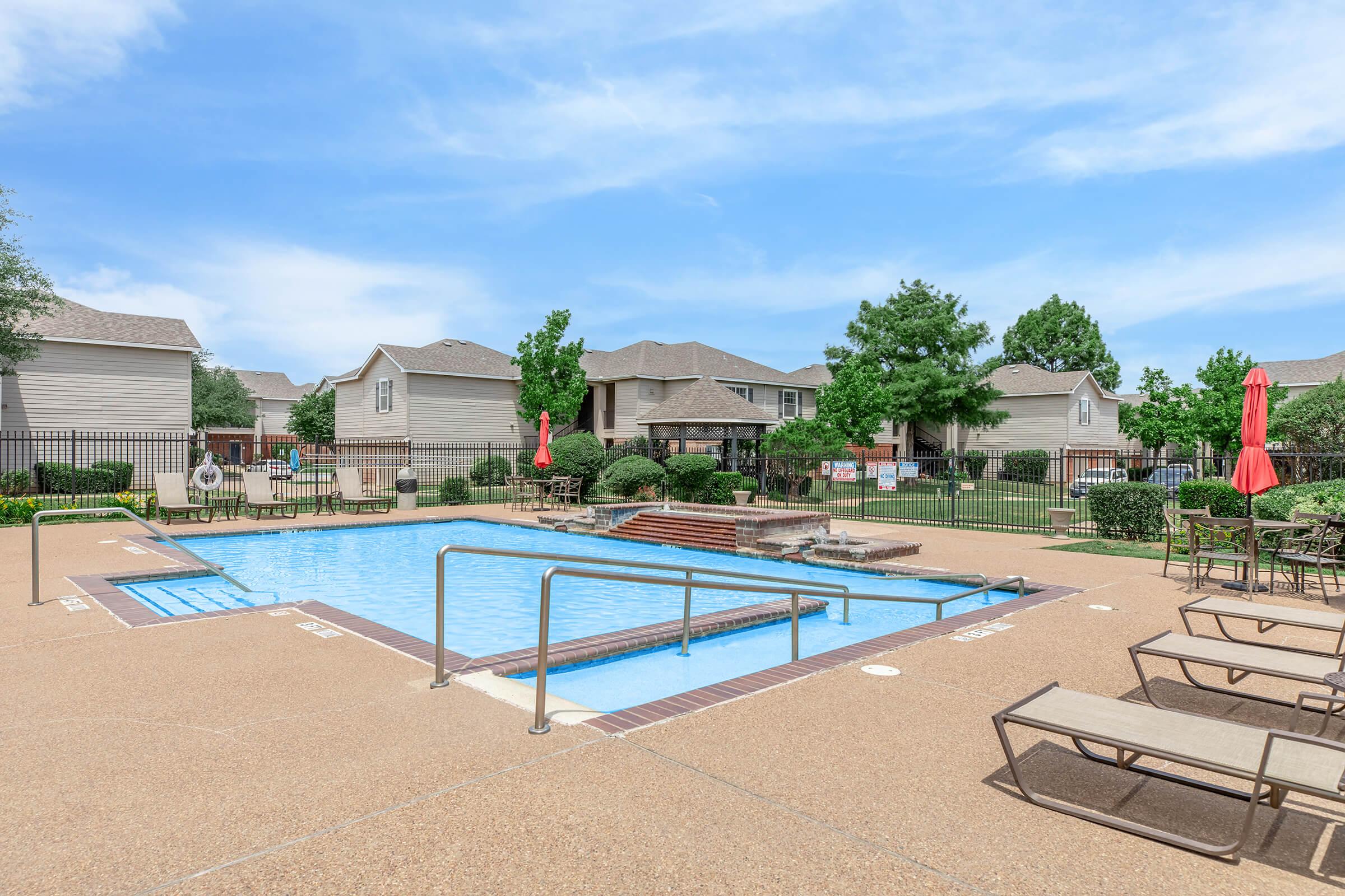 A swimming pool surrounded by lounge chairs and umbrellas. The pool area is fenced and features green landscaping with trees and shrubs. In the background, there are residential buildings, creating a relaxed outdoor atmosphere on a sunny day.