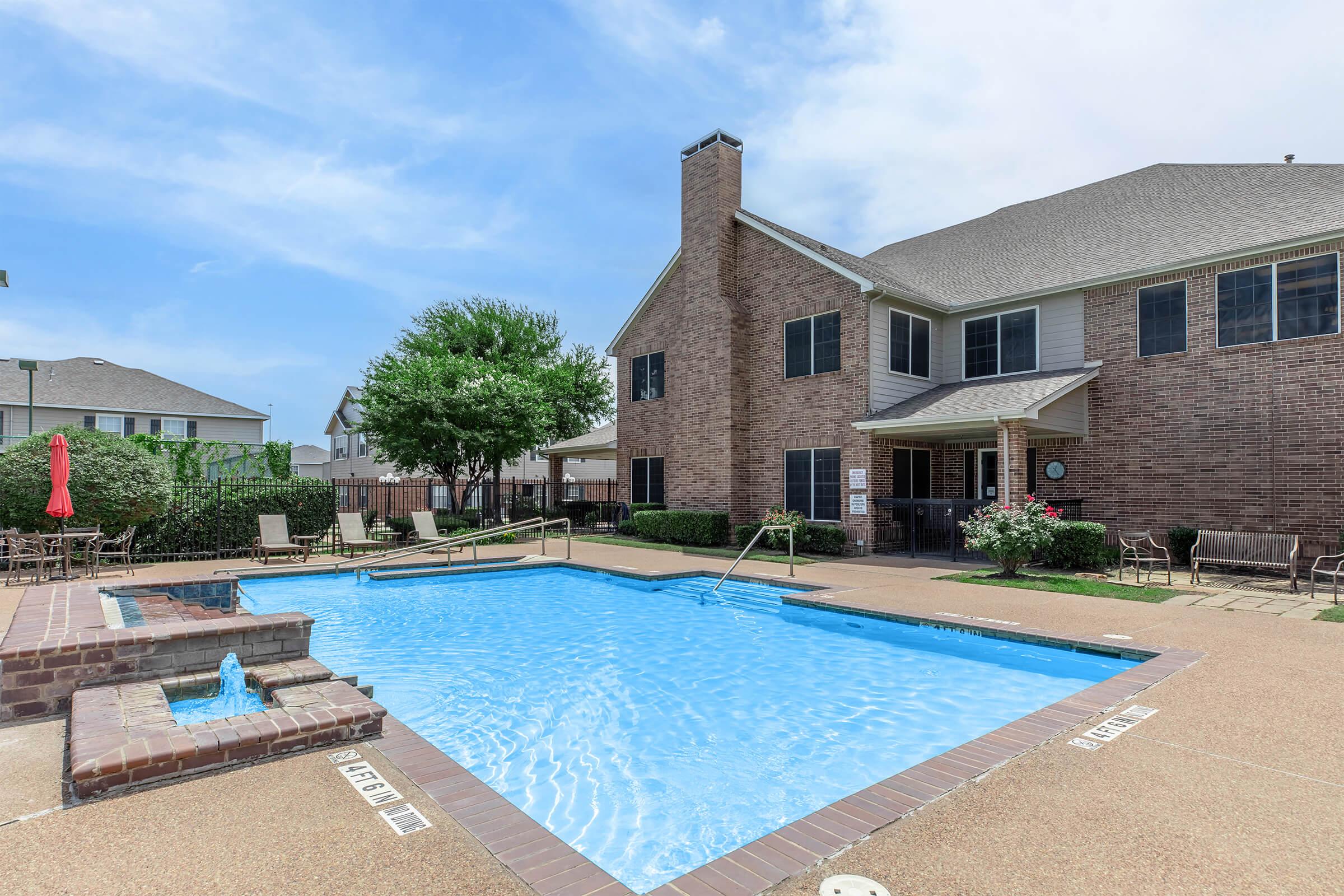 A clear blue swimming pool surrounded by a patio area, featuring lounge chairs and a hot tub. In the background, a two-story brick building with large windows and a well-maintained landscape. The sky is bright with a few clouds, creating a sunny and inviting atmosphere.