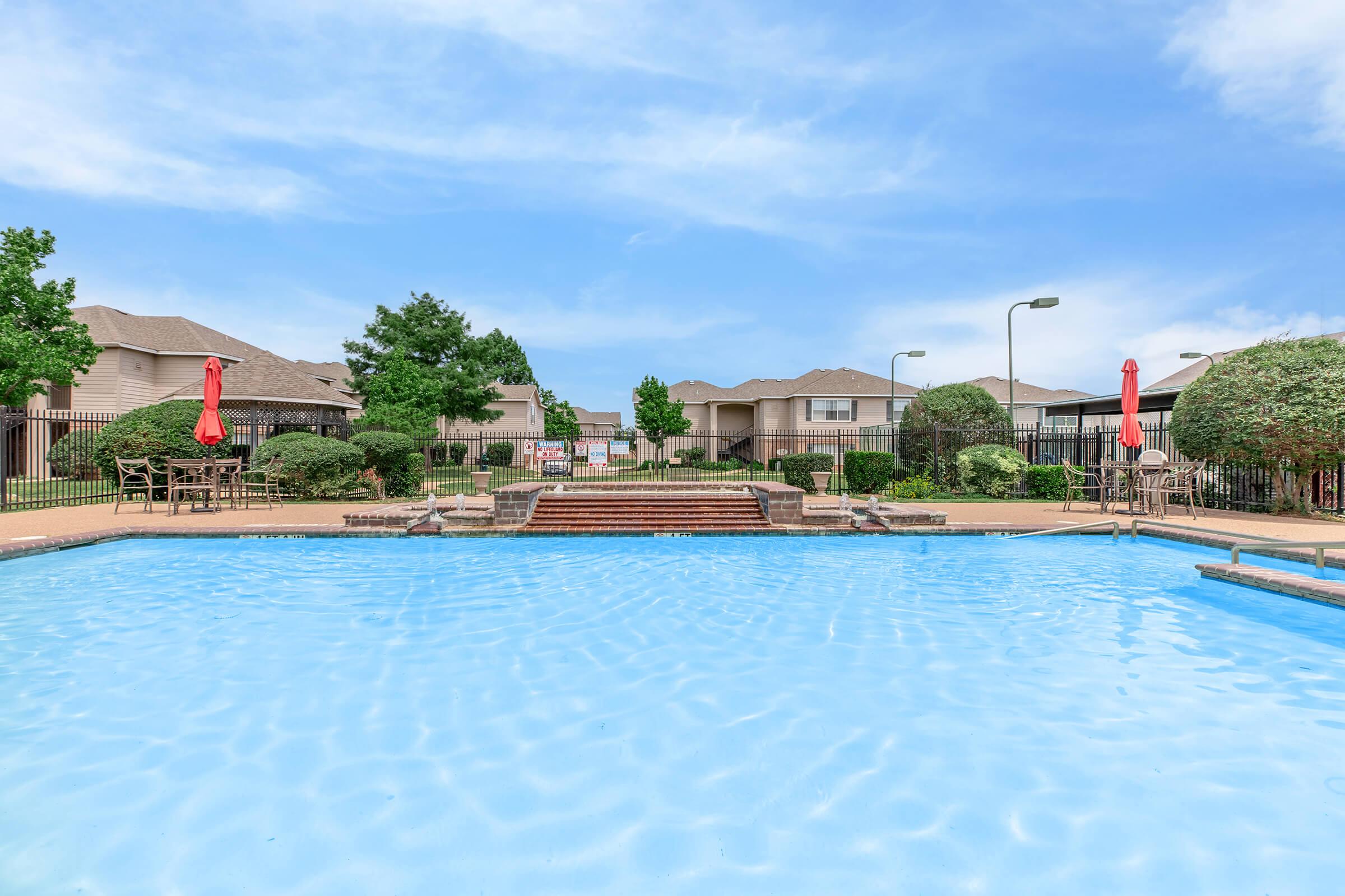 A clear blue swimming pool in a landscaped area, surrounded by green shrubs and trees. There are red umbrellas and patio furniture visible. Residential buildings in the background and a blue sky above create a serene outdoor setting.