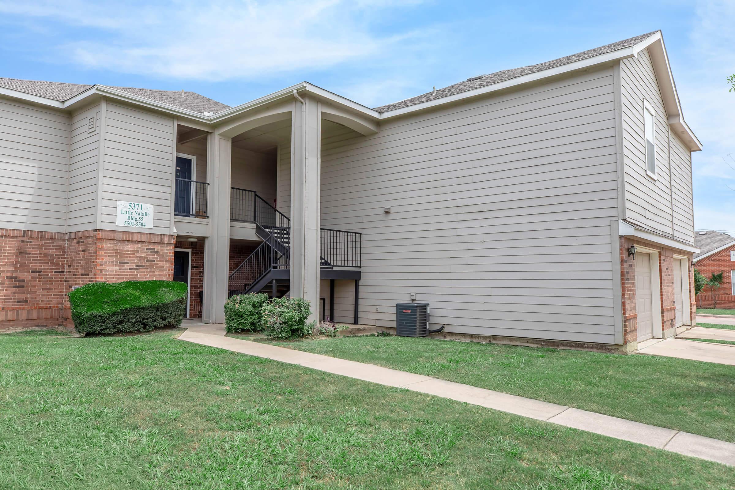 Exterior view of a multi-unit residential building with light gray siding and brick accents. A metal staircase leads to the second floor, surrounded by landscaped grass and shrubs. The setting is suburban, under a clear blue sky. Pathways provide access to the building entrance.