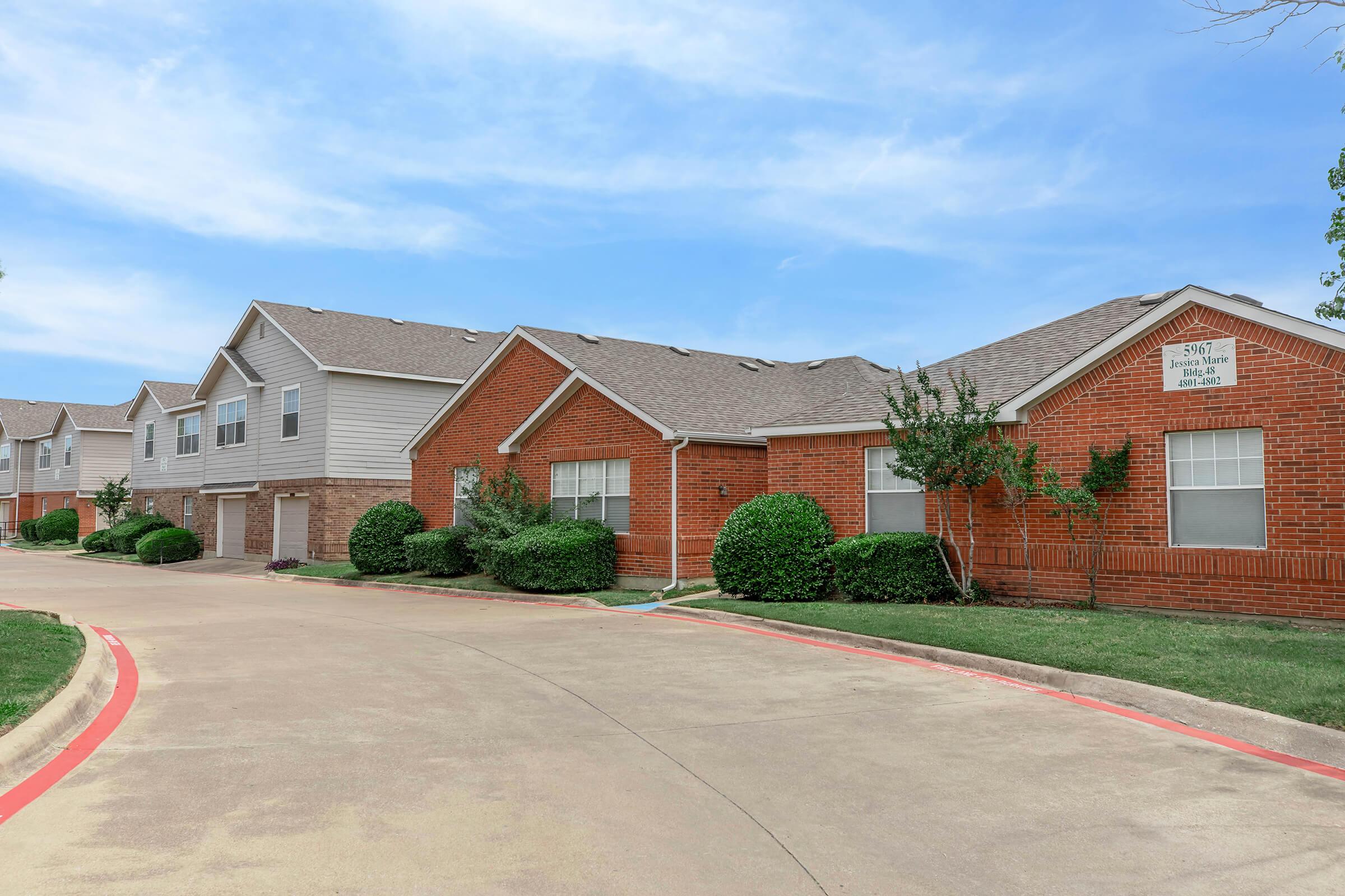 A view of residential buildings with brick facades and gray siding, set along a paved driveway. The landscape features well-kept shrubbery and grass, under a blue sky with scattered clouds. The setting appears to be a quiet apartment complex or neighborhood.