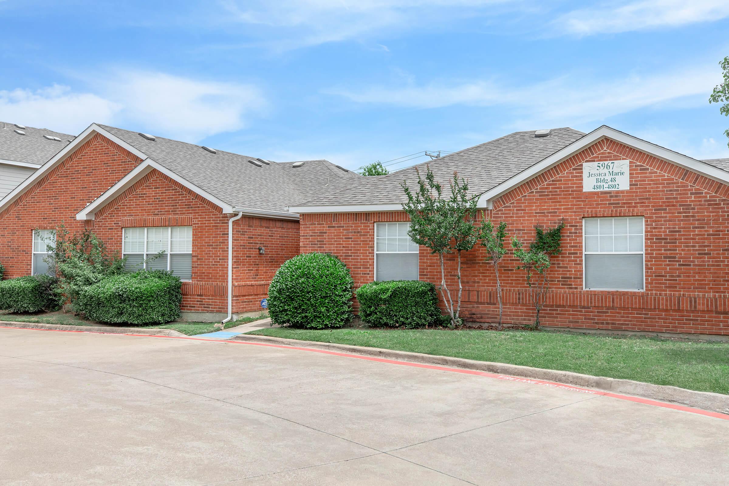 Single-story red brick residential building with a sloped gray roof. It features multiple windows and manicured bushes in front. A paved driveway is visible, leading to the entrance, which has a sign with an address displayed. The surrounding area is green with grass and small trees under a blue sky.