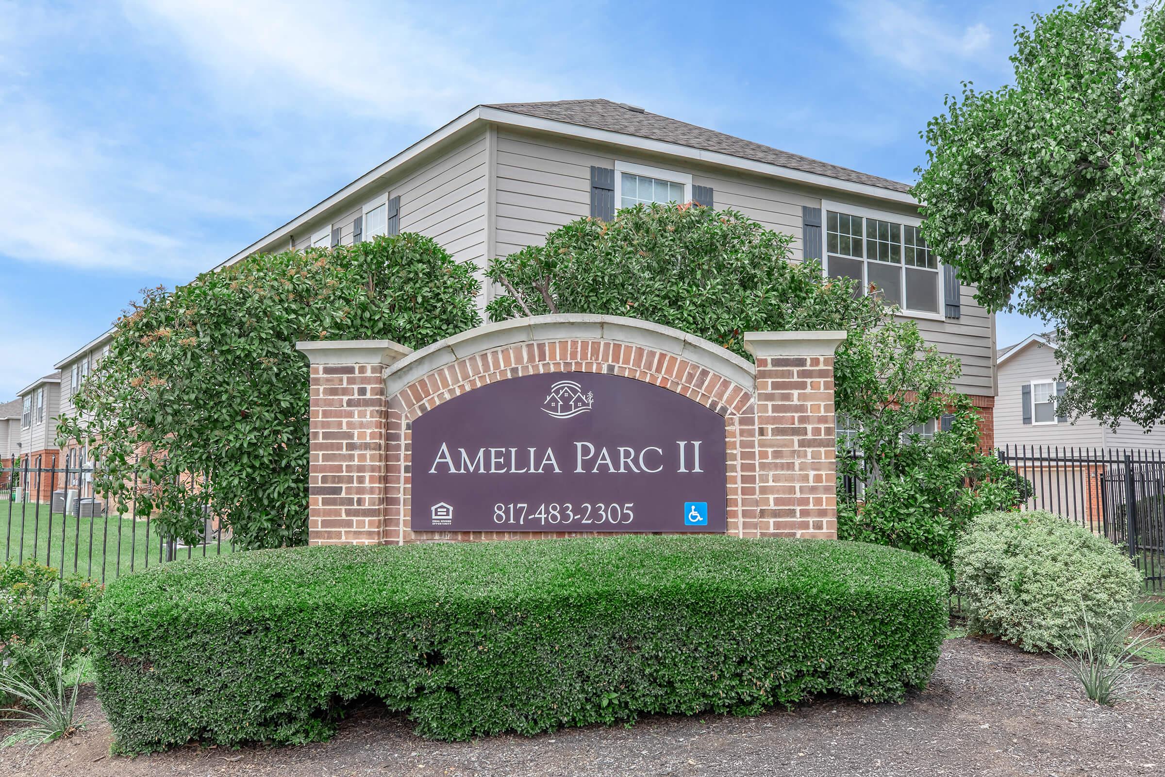 Sign for Amelia Parc II, a residential community, featuring a brick structure with landscaping. The sign includes the community name, contact number, and accessibility symbol. In the background, a modern building and clear blue sky are visible.