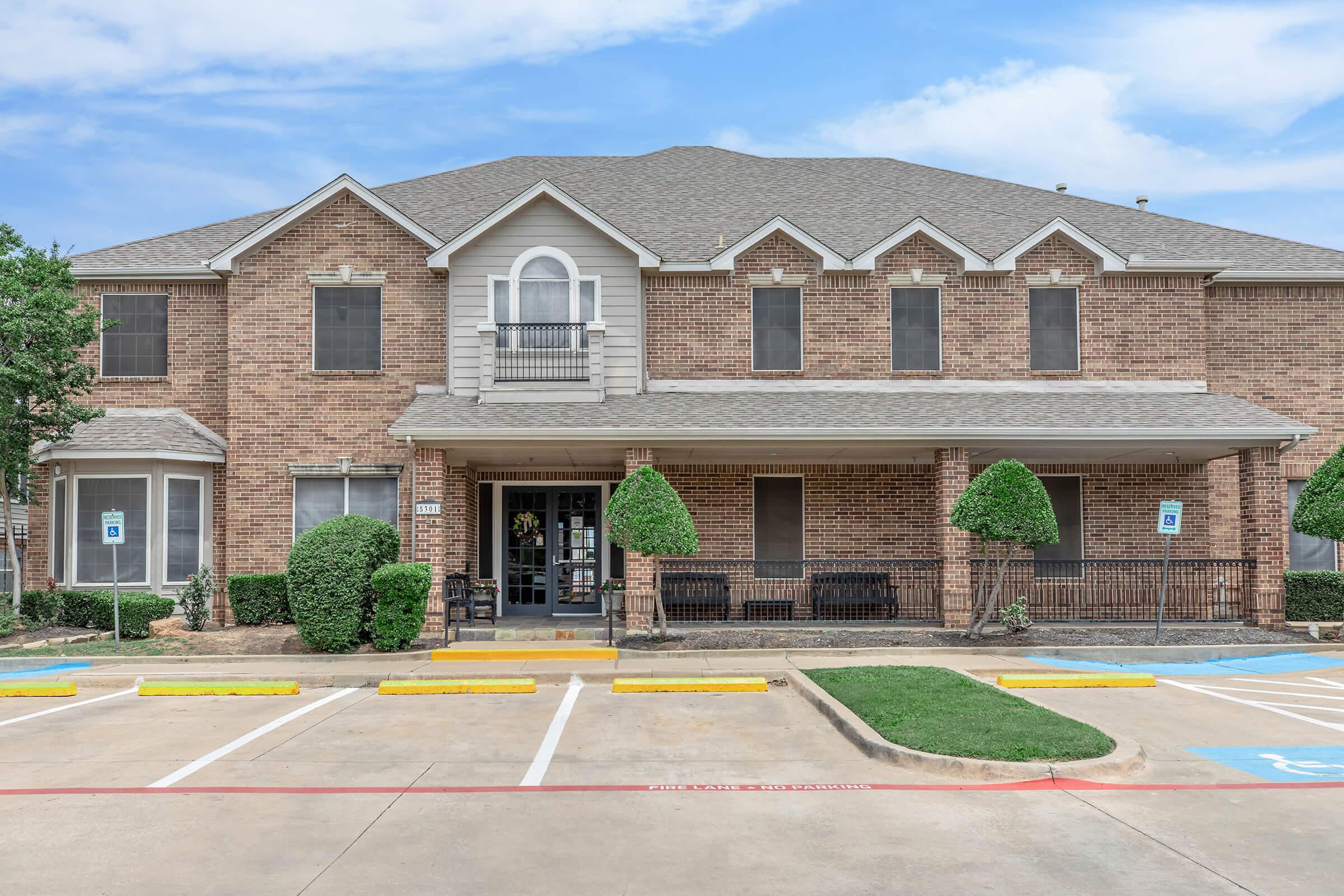 A brick building with two stories featuring large windows and a covered entrance. The front has manicured shrubs and a paved parking area with designated spots. The sky is partly cloudy, adding to the pleasant appearance of the property.