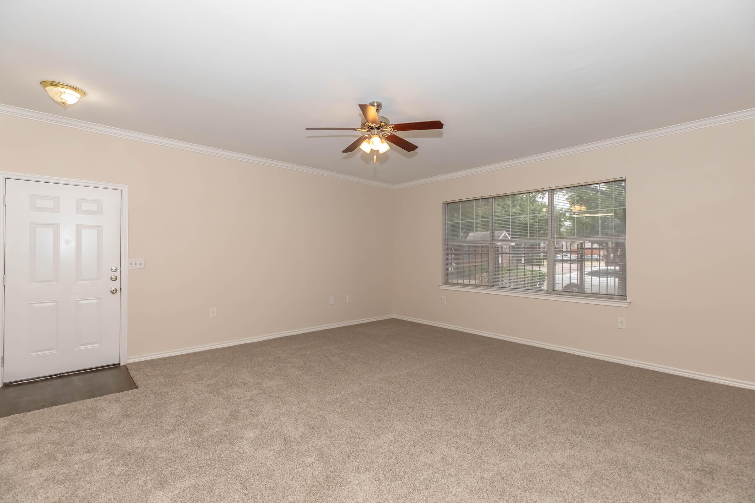 A spacious, light-colored living room with beige walls and carpet. The room features a ceiling fan and a large window with blinds, allowing natural light to enter. A front door is visible on the left side, leading to the exterior. The space is empty, offering a clean and uncluttered look.