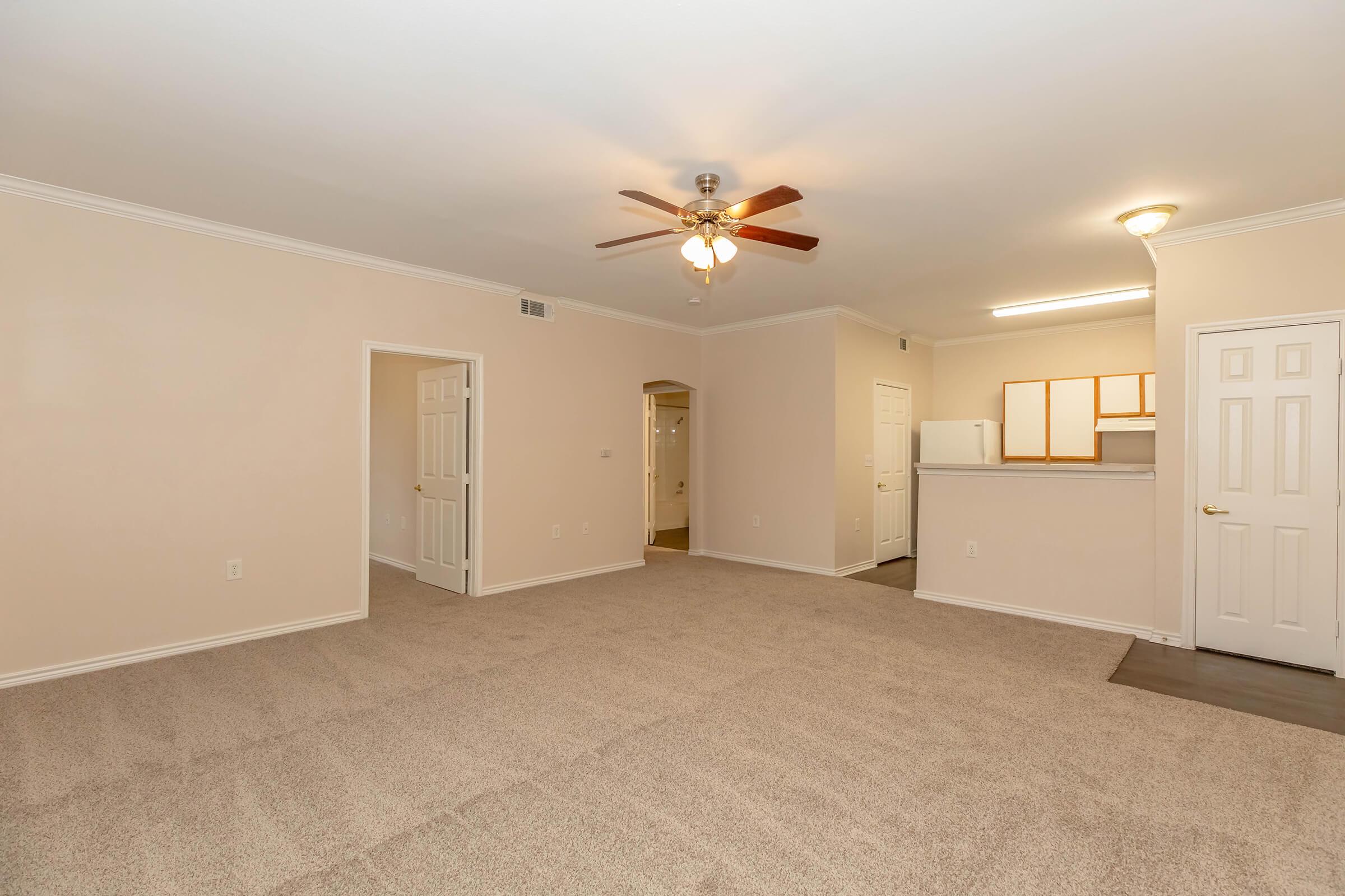 Spacious and well-lit living area featuring light beige walls and soft carpet. A ceiling fan hangs in the center, and open doorways lead to adjacent rooms. To the right, a kitchen area is visible with hardwood flooring. Natural light floods the space, creating a welcoming atmosphere.