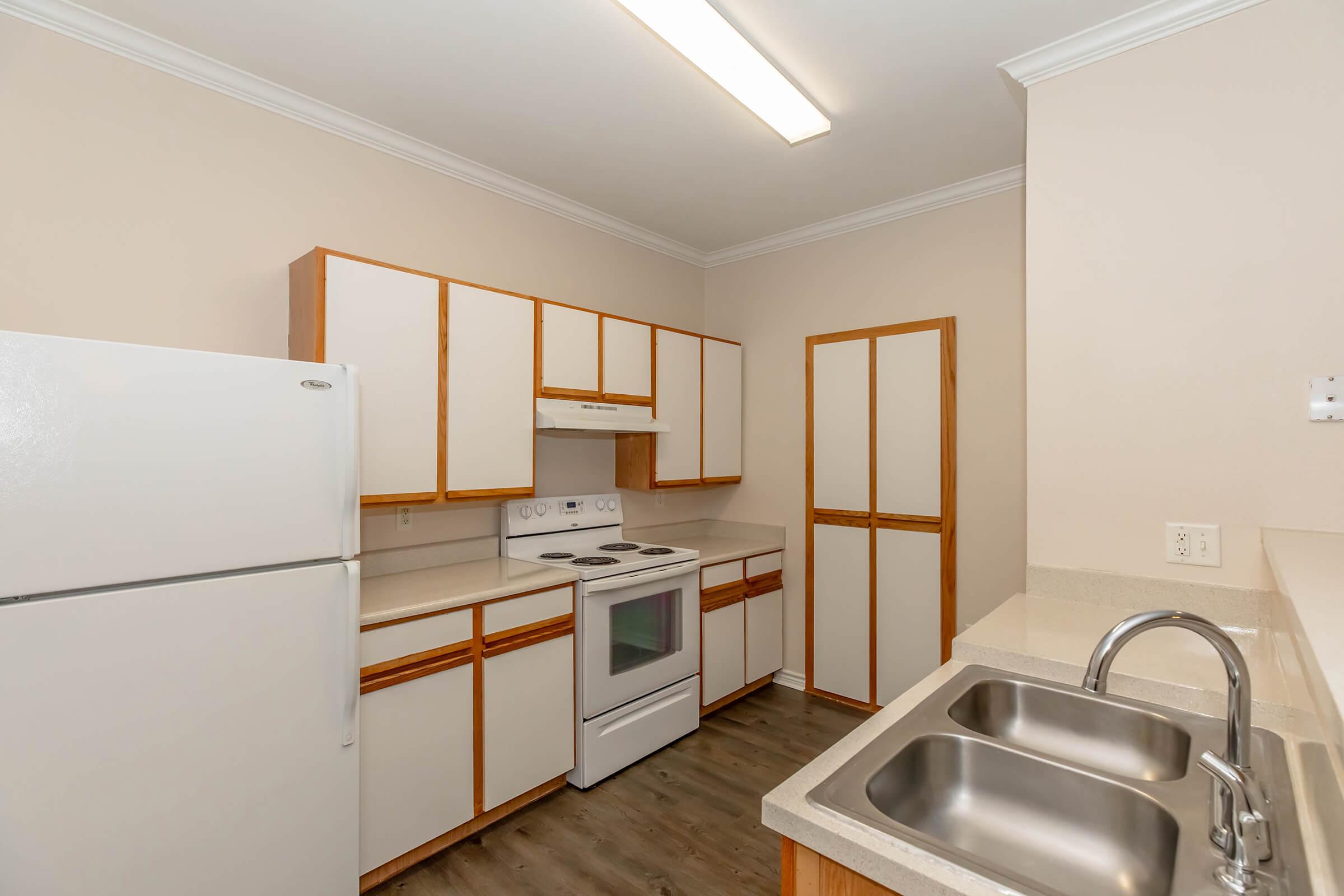A clean, modern kitchen featuring white cabinets with wooden accents, a white refrigerator, an oven with a stovetop, and a double sink. The room has beige walls, light-colored countertops, and ample lighting from a ceiling fixture, creating a bright and inviting atmosphere.