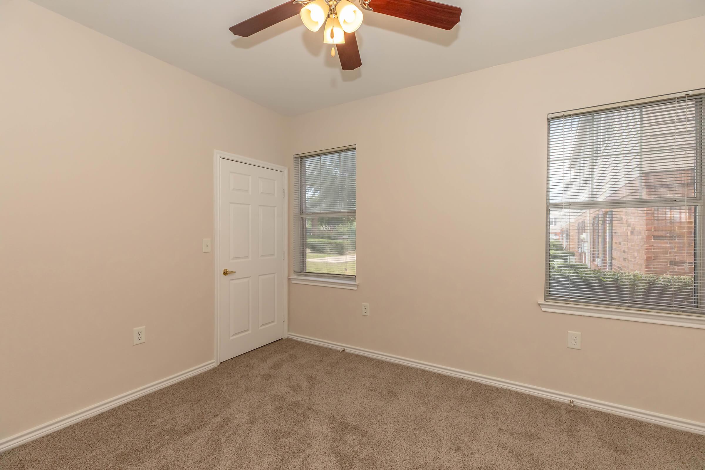 A light-colored room with beige walls and a carpeted floor. It features two windows with blinds, a ceiling fan with lights, and a white door. The space is empty, providing an uncluttered and bright atmosphere.