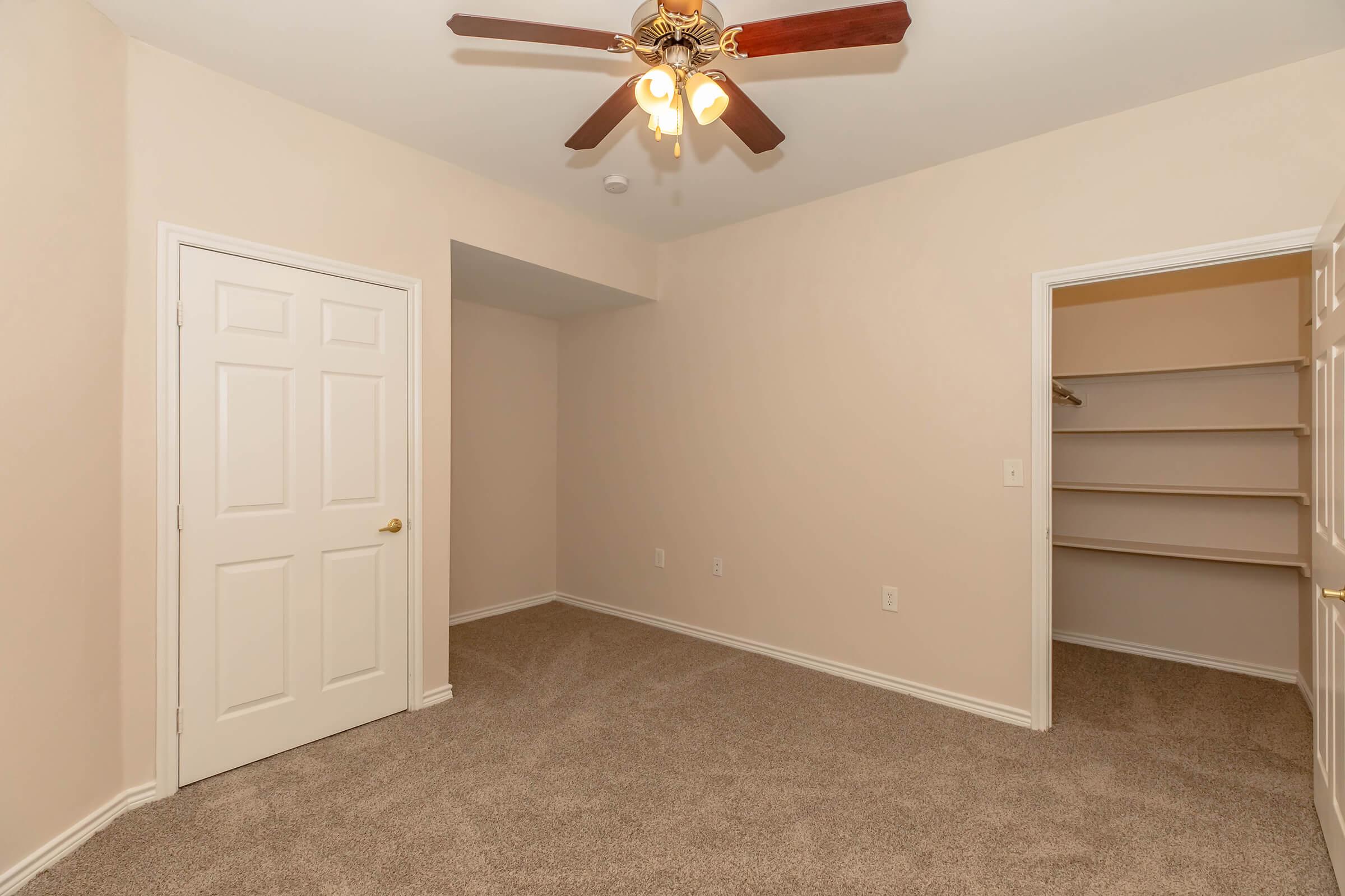 A well-lit bedroom featuring a ceiling fan with light fixtures, beige walls, and carpeted flooring. It includes a closed door leading to an adjacent closet with shelving. The room appears spacious and has a neutral color palette, creating a warm and inviting atmosphere.