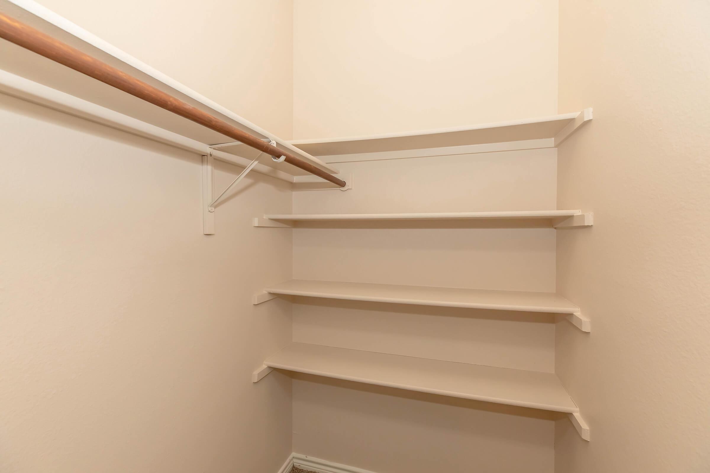 Empty closet with beige walls and wooden clothing rod. Shelves are neatly arranged on one side, providing storage space. The overall appearance is clean and simple, suitable for organizing clothes or other items. Natural light enhances the spacious feel of the closet.