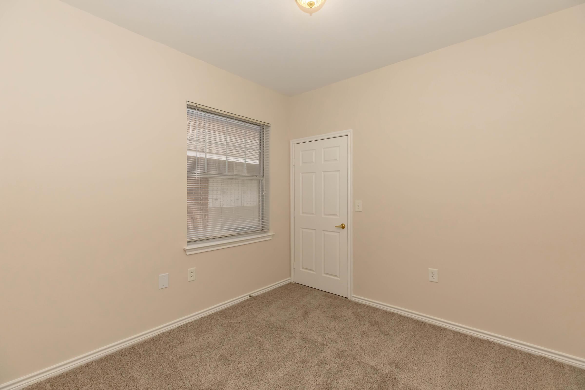 A small, neutral-colored room with beige walls and light brown carpet. There is a window with blinds allowing natural light, and a plain white door. The room appears empty, giving it a spacious and minimalist feel.