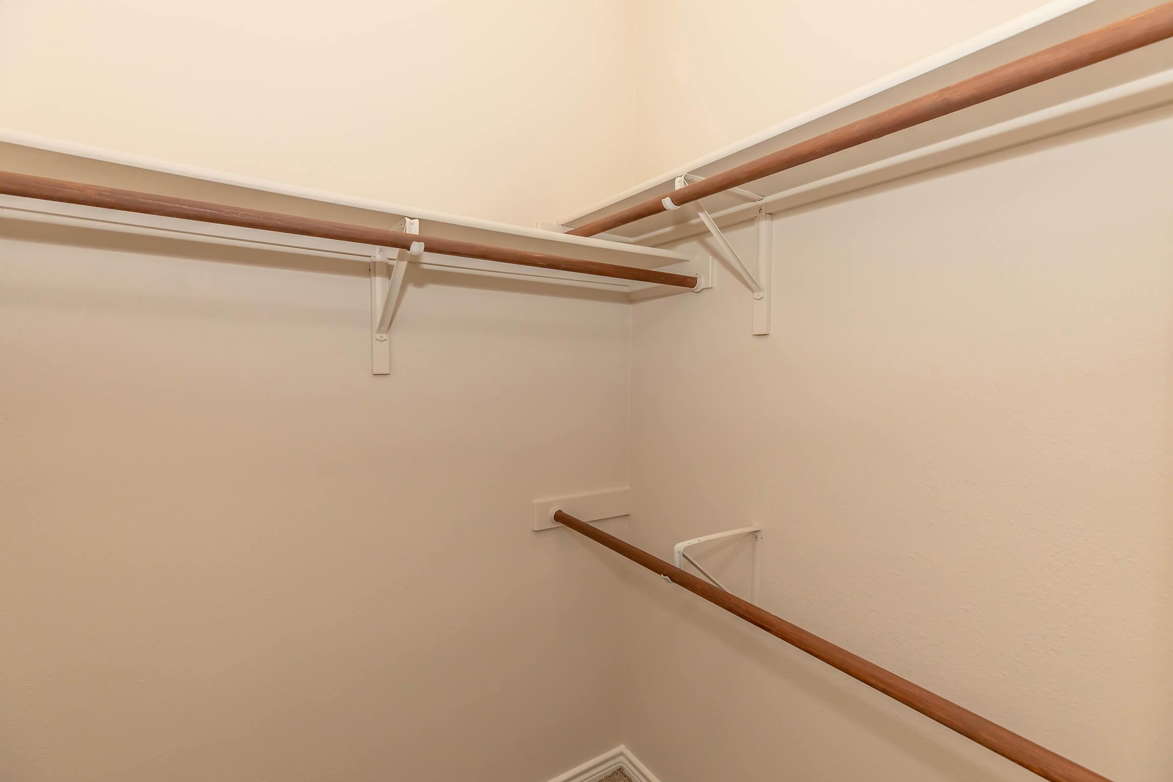 Empty closet with two wooden hanging rods mounted on white brackets against a light-colored wall. The space is unadorned and has no shelving or additional storage items visible.