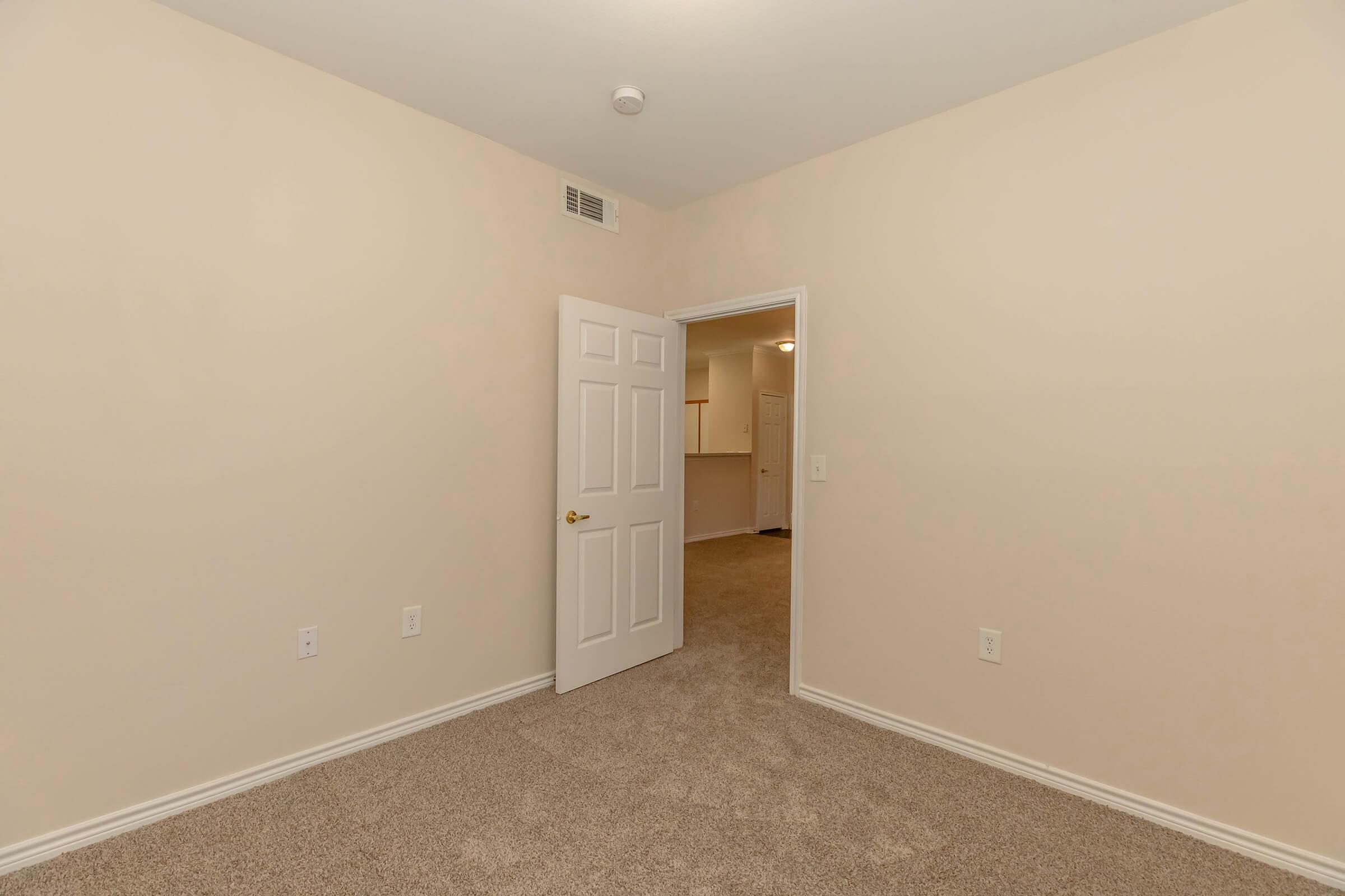Interior view of a small, empty beige room with carpeted flooring. A closed white door is visible on the left, leading to another space. The walls are plain and unadorned, offering a neutral backdrop. There is a light fixture on the ceiling, and a doorway is seen in the background.